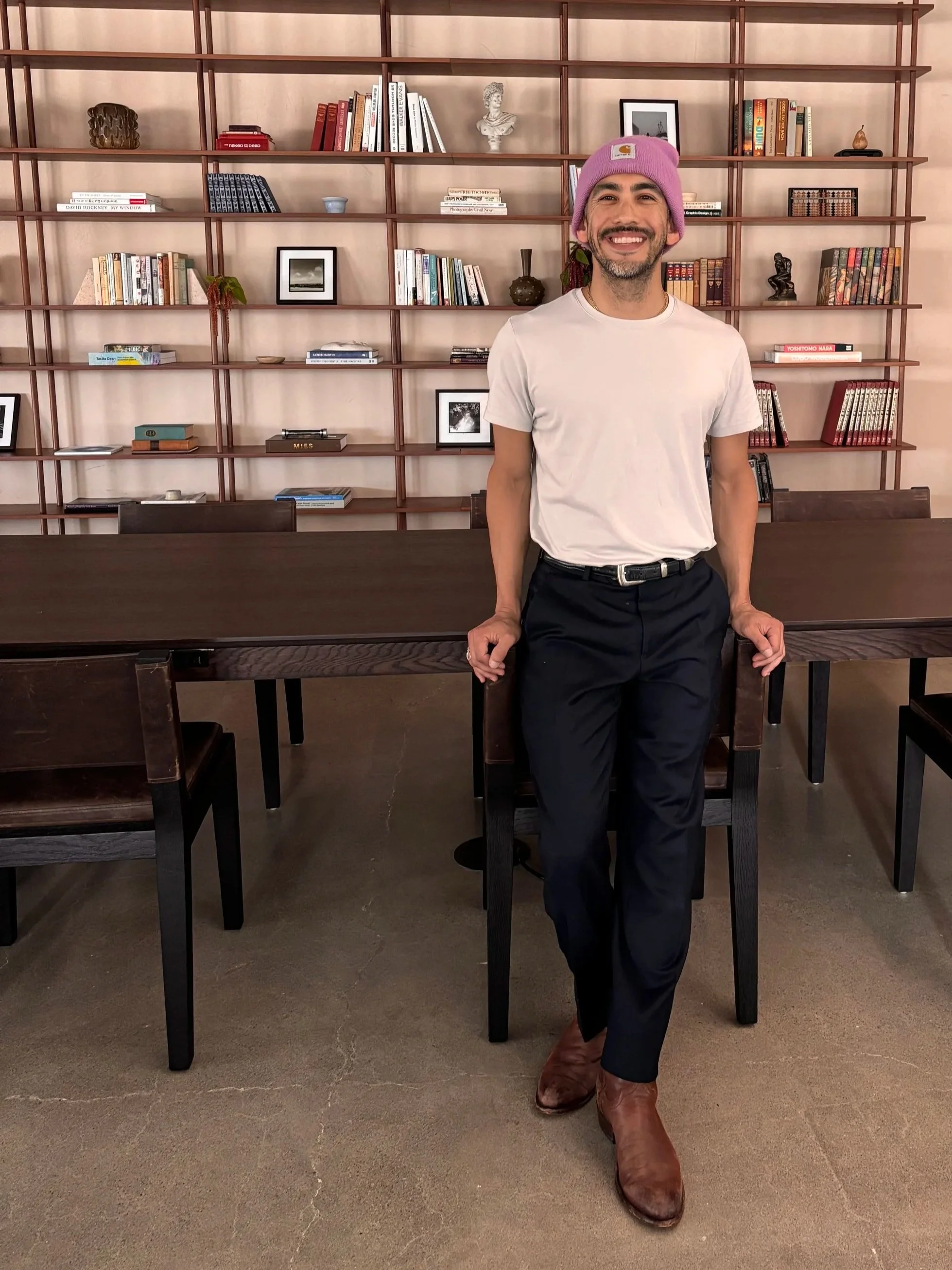 A smiling man with a mustache, wearing a pink beanie, a white t-shirt, black pants, and brown boots, sitting in a modern room with a large wooden bookshelf in the background filled with books and decorative items. Styled by Adrian Jacob Creative.