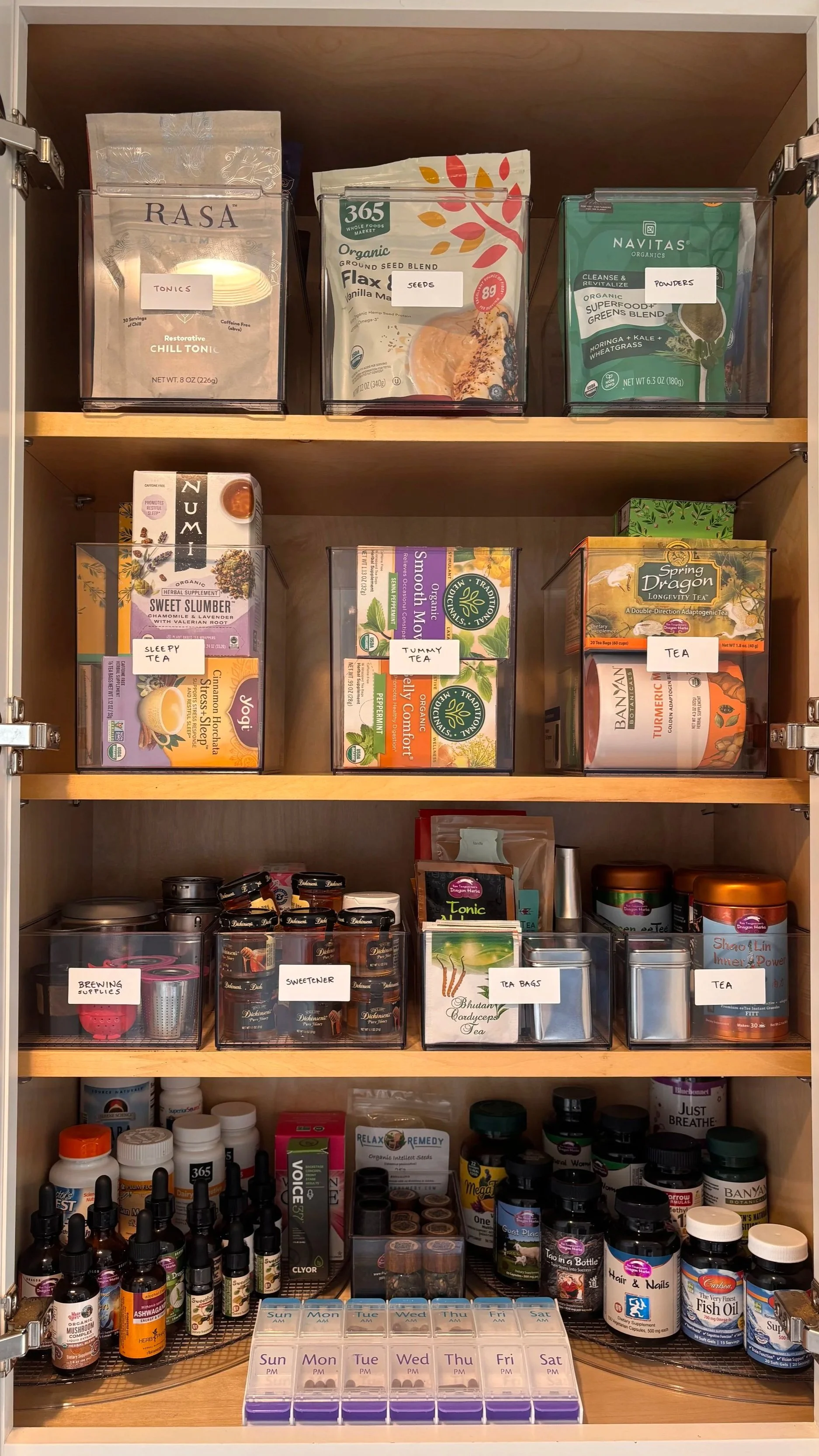 A wooden shelf with various health and wellness products, including teas, supplements, tinctures, and labeled containers, organized in plastic bins. Kitchen organized by Adrian Jacob Creative