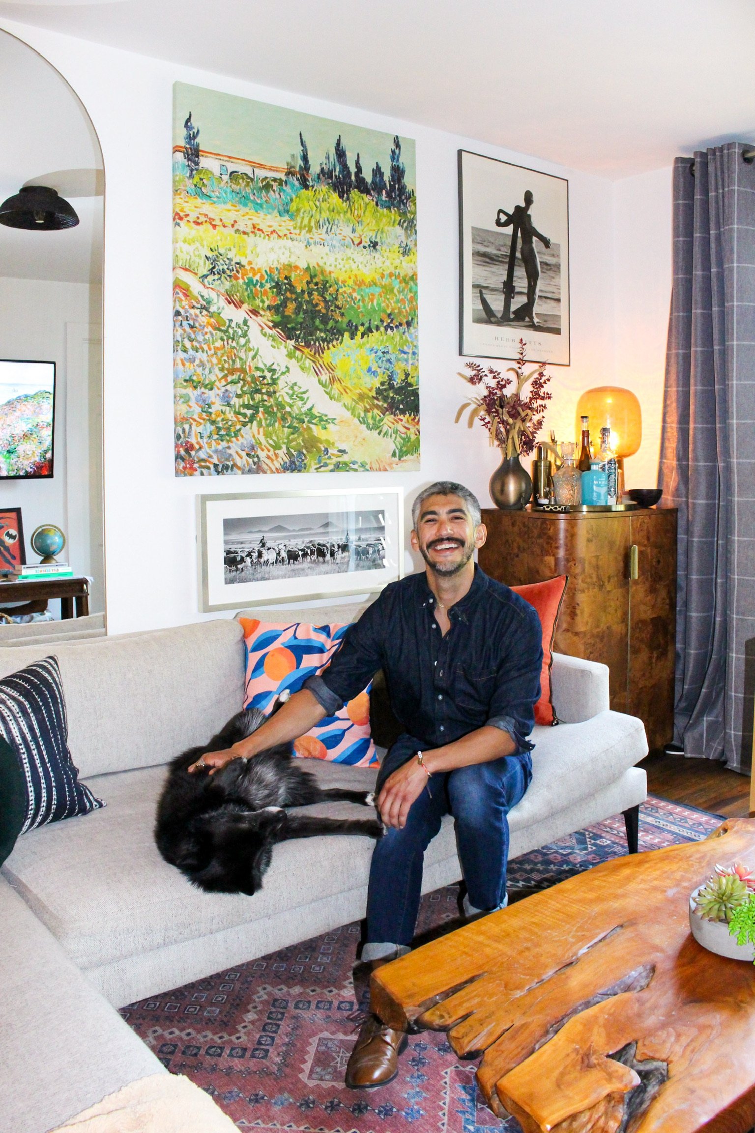A man with gray hair and a beard smiling while sitting on a sofa next to a black dog with white markings, in a living room with colorful artwork on the walls, a wooden coffee table, a sideboard with decorative bottles and a glowing lamp, and a patterned rug.