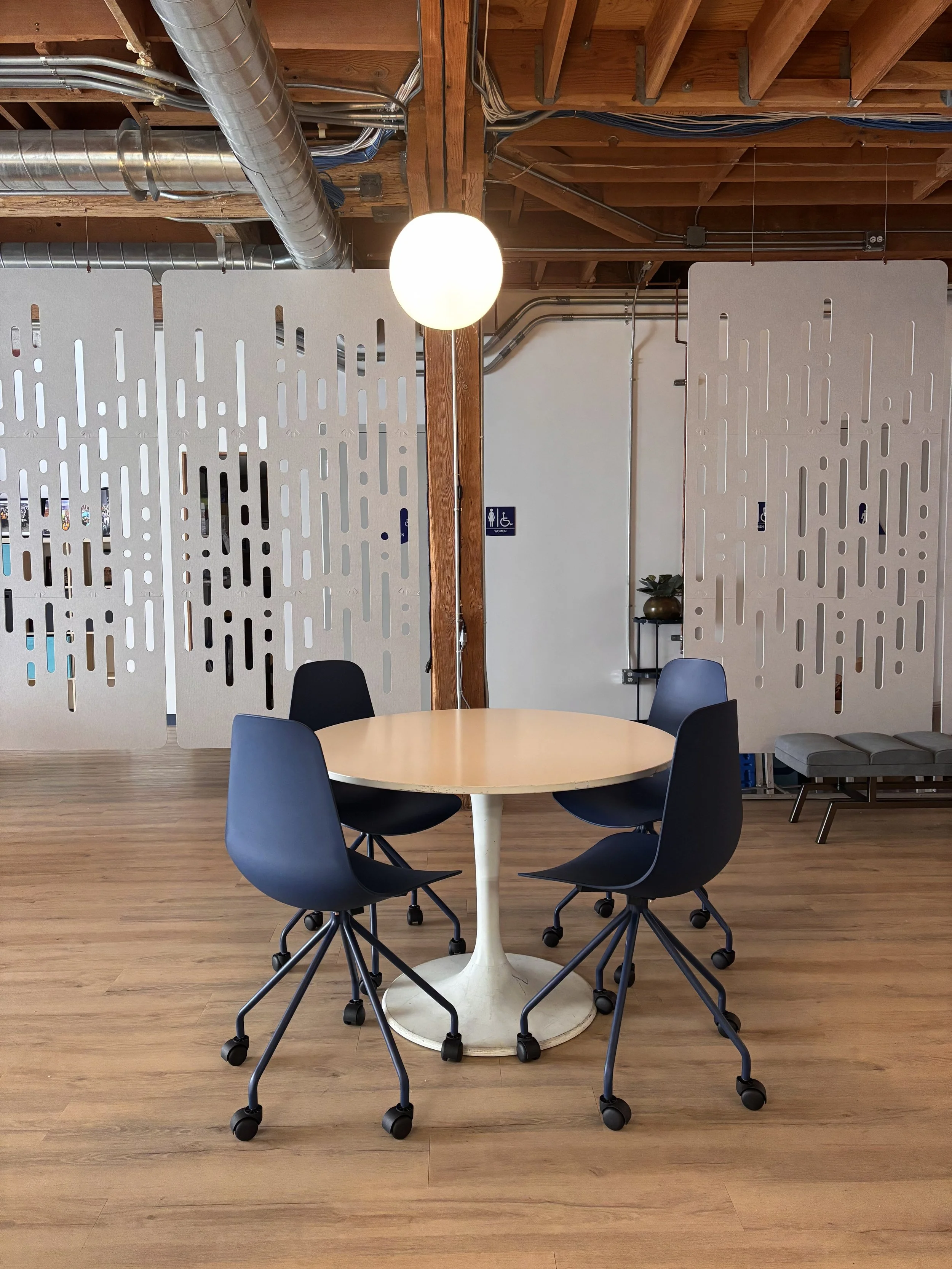 A round table with navy chairs on caster wheels, in an office with wooden flooring, a wooden ceiling with exposed beams, ductwork, and partition panels, as well as a potted plant and a bench in the background. Styled by Adrian Jacob Creative.