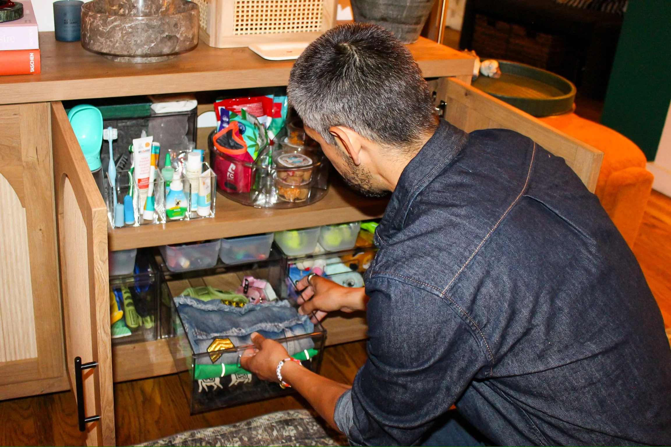 A person with short dark hair wearing a denim shirt (Adrian Jacob) is organizing items in a wooden drawer with multiple transparent compartments filled with various pet supplies.