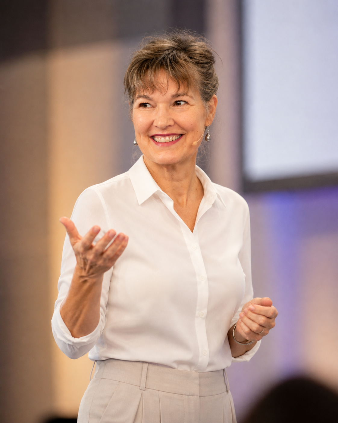 A woman with short, brown hair smiling and gesturing with her right hand, wearing pearl earrings, a white button-up shirt, and beige pants, speaking in an indoor setting with a blurred background.