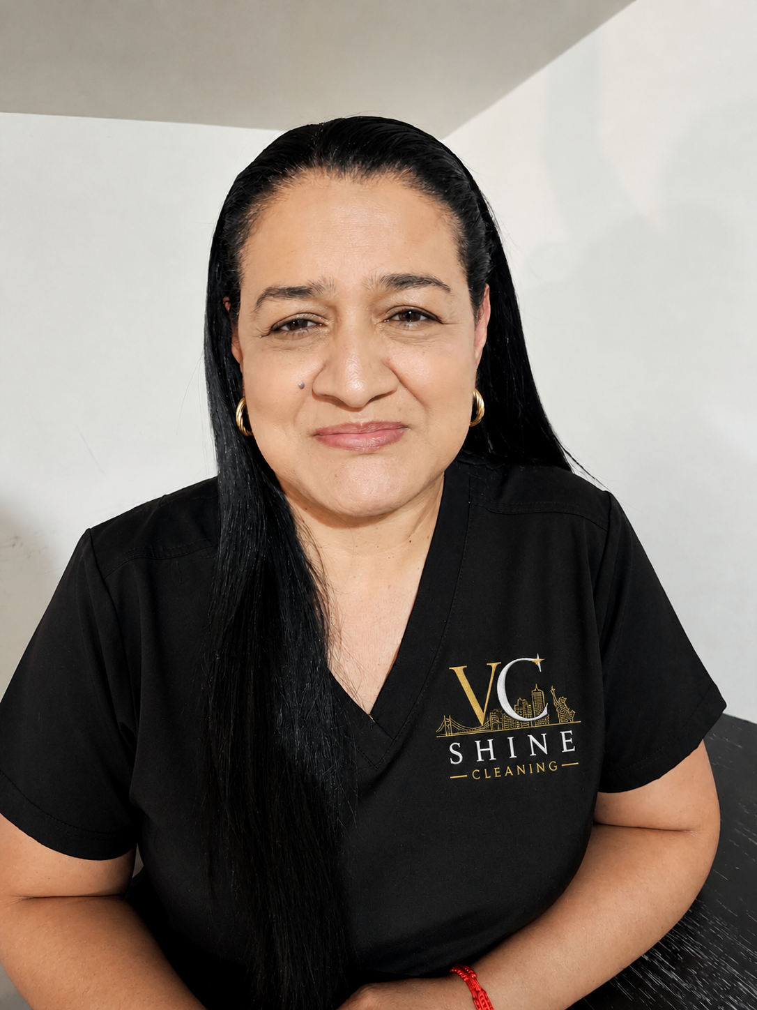A woman with long black hair, wearing gold hoop earrings, a black shirt with the words 'VC Shine Cleaning' and a graphic of city landmarks, sitting at a table in a room with plain white walls.