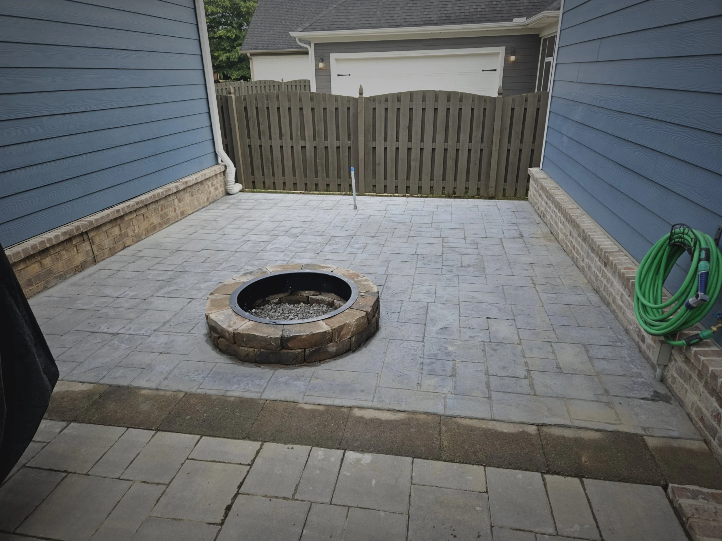 Patio with stone pavers, fire pit, green garden hose on the right, blue house siding on the left, and a wooden fence with a gate in the background.