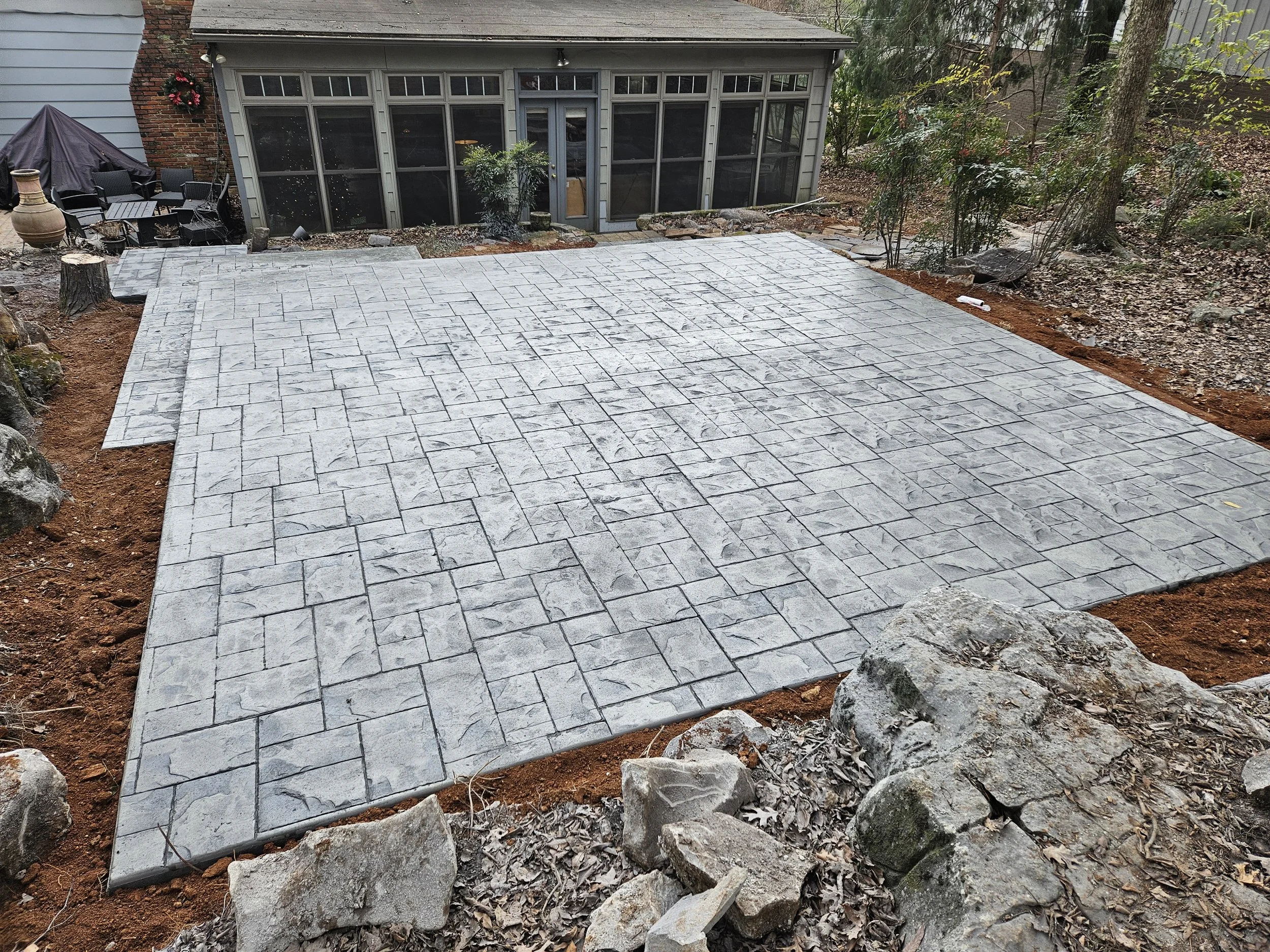 A newly paved stone patio with gray interlocking stones, surrounded by dirt and rocks, in a backyard with a screened porch, outdoor furniture, trees, and plants.