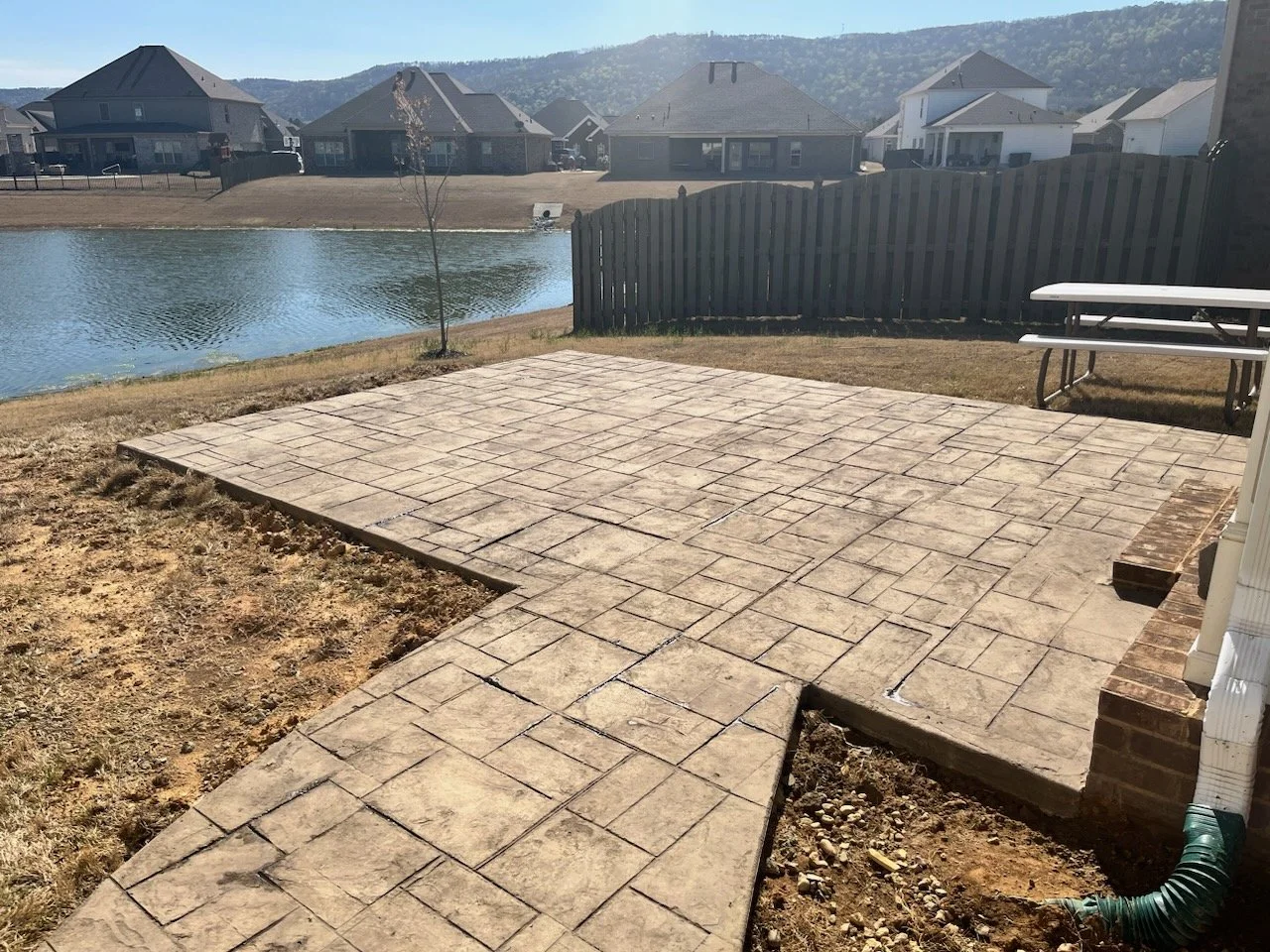 Backyard patio with brick and concrete pavers, small pond, fencing, nearby houses, and hills in the background.