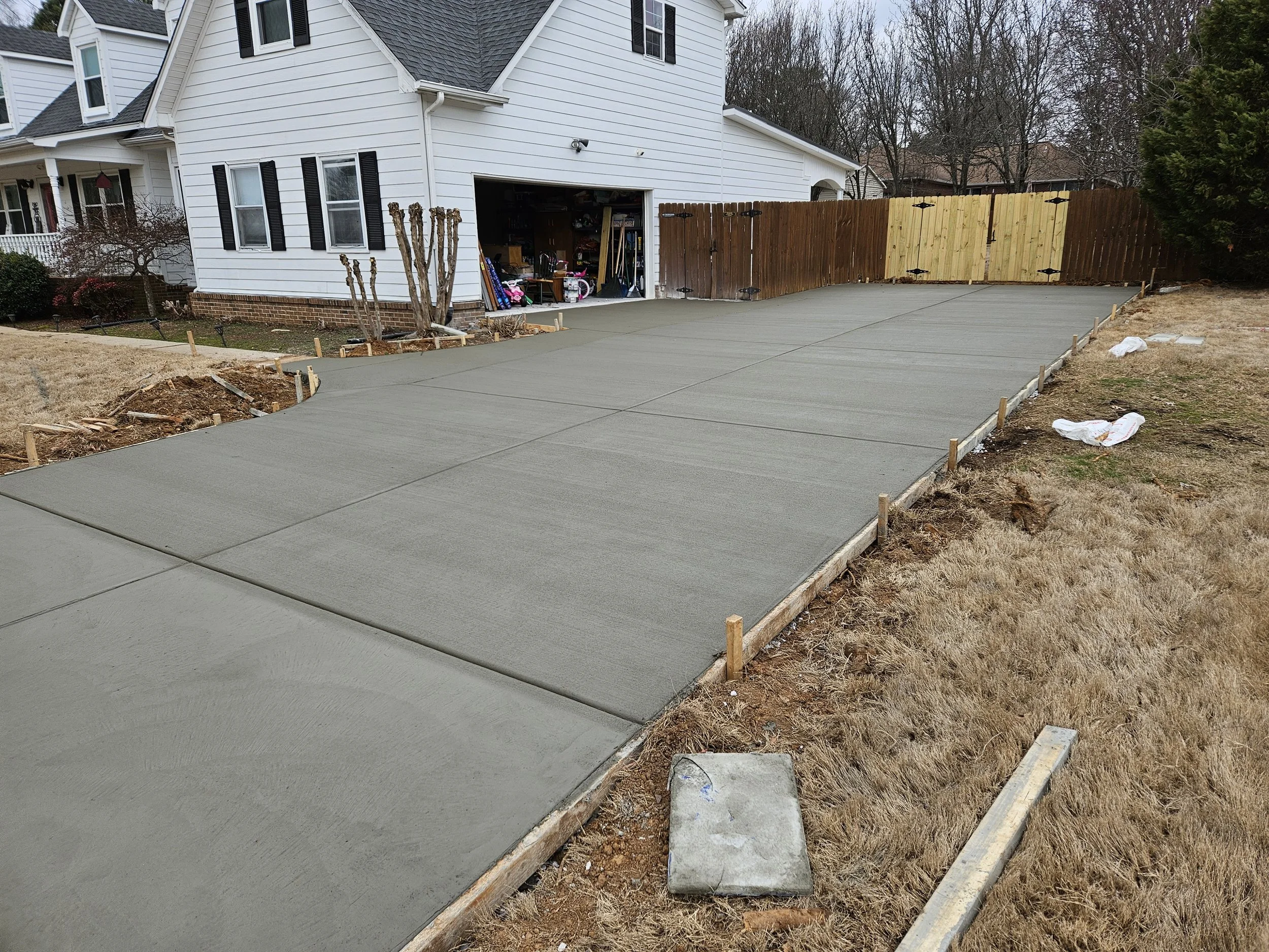 A house with an attached garage under construction, with a freshly poured concrete driveway. The garage is open, revealing tools and storage within. Surrounding the driveway, there is bare soil and construction materials, with a wooden fence partiall