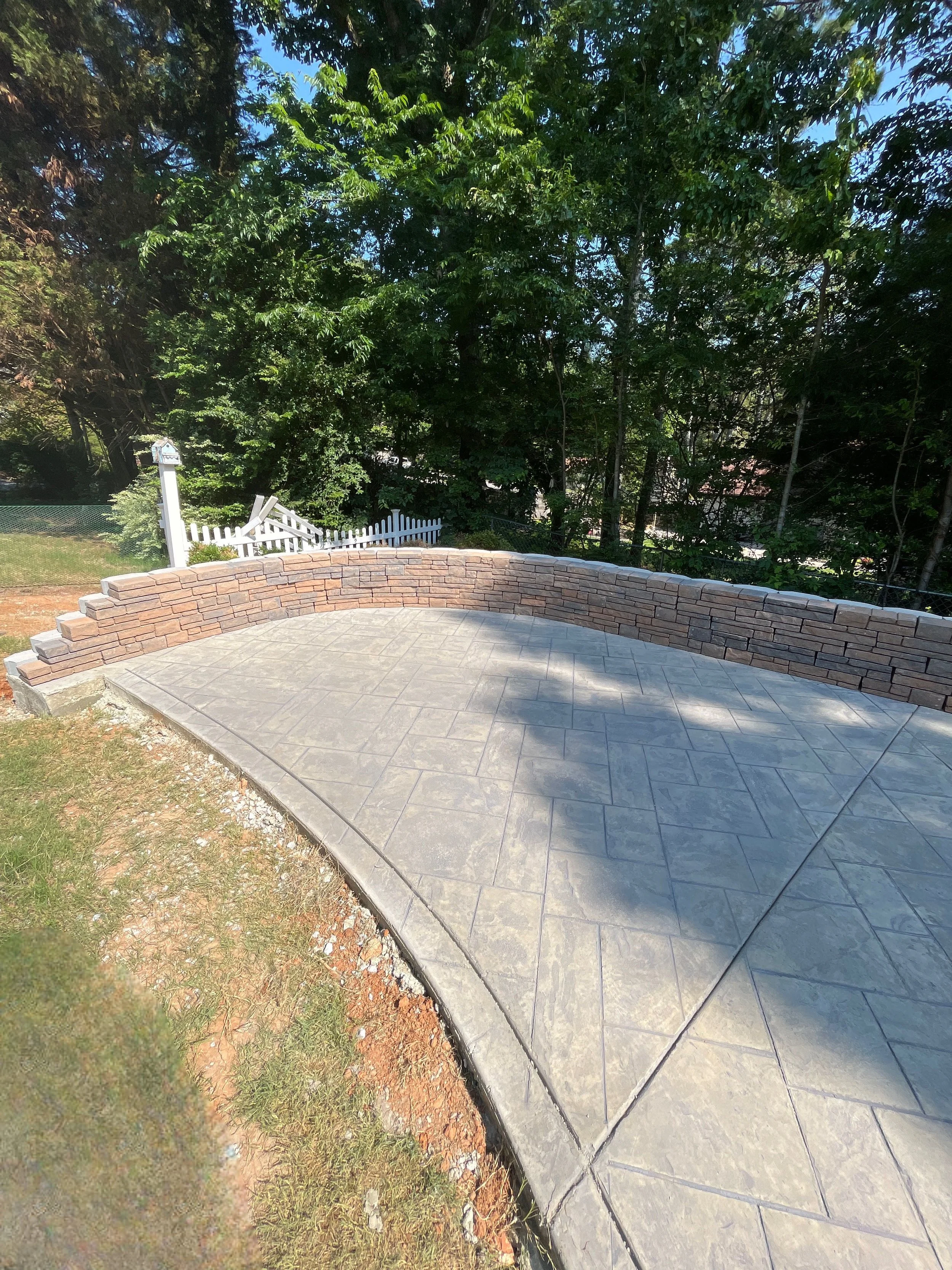 A curved brick and concrete patio with a brick border, adjacent to a grassy area and a dirt patch, surrounded by trees and a white picket fence in the background.