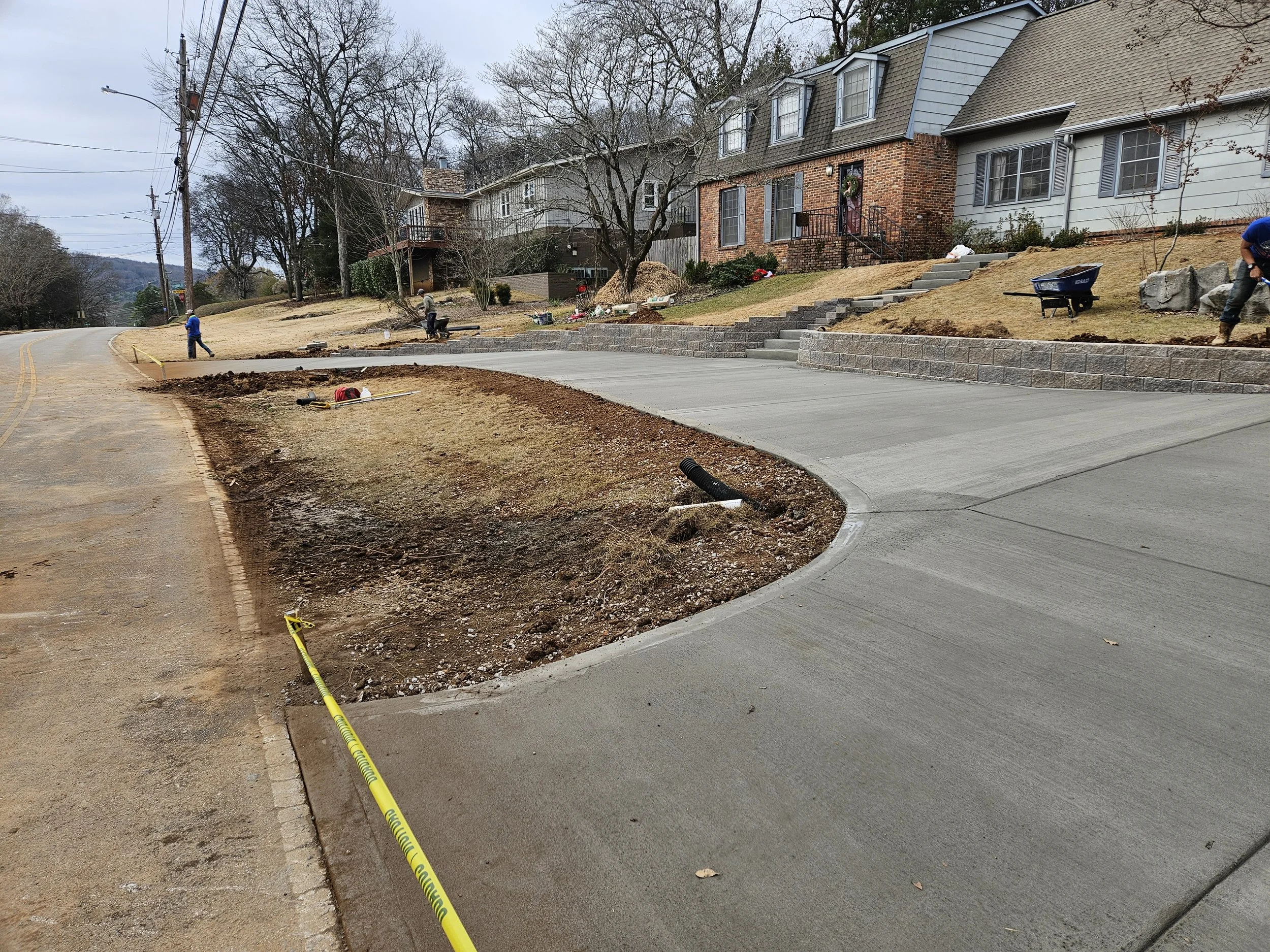 Construction workers are building a retaining wall along a sloped residential yard, with stairs leading up to houses. There is construction tape and tools on the ground.