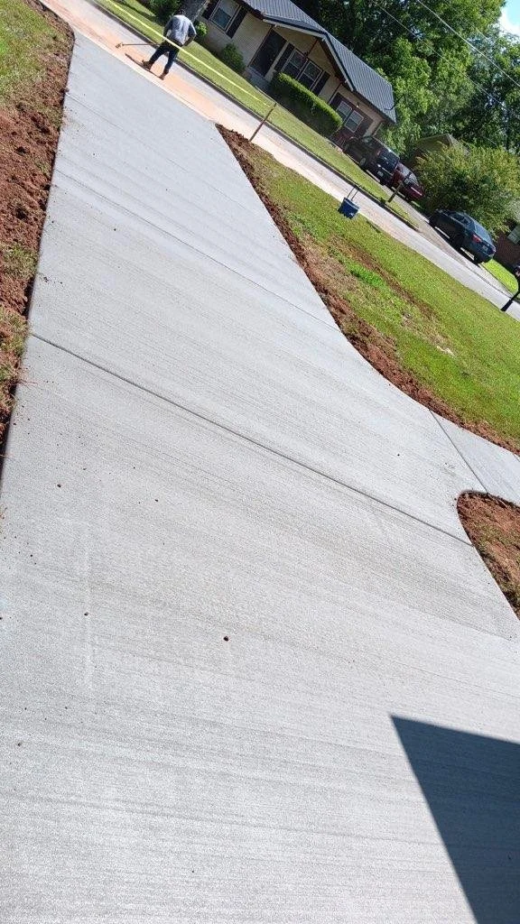 Newly poured concrete sidewalk with a person working on it, in a residential neighborhood with houses, trees, and parked cars visible in the background.