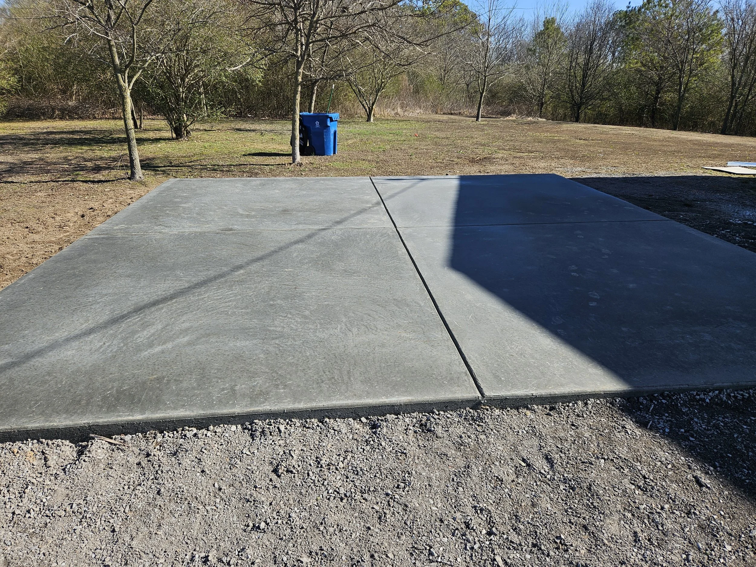 A concrete driveway next to a grassy yard with trees and a blue recycling bin. Shadows cast from the left side, with a clear blue sky in the background.
