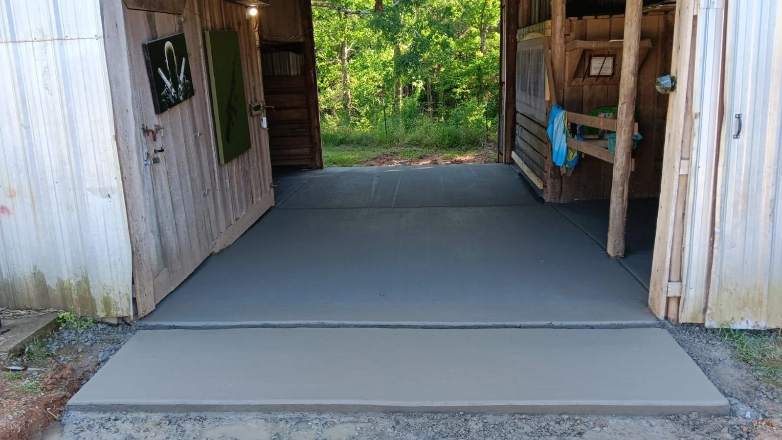 Freshly poured concrete landing in the open entrance of a rustic wooden shed with outdoor greenery visible in the background.