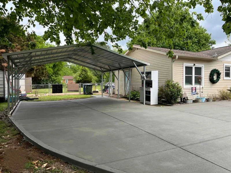 Concrete driveway with a metal carport, attached to a beige house with a wreath on the wall.