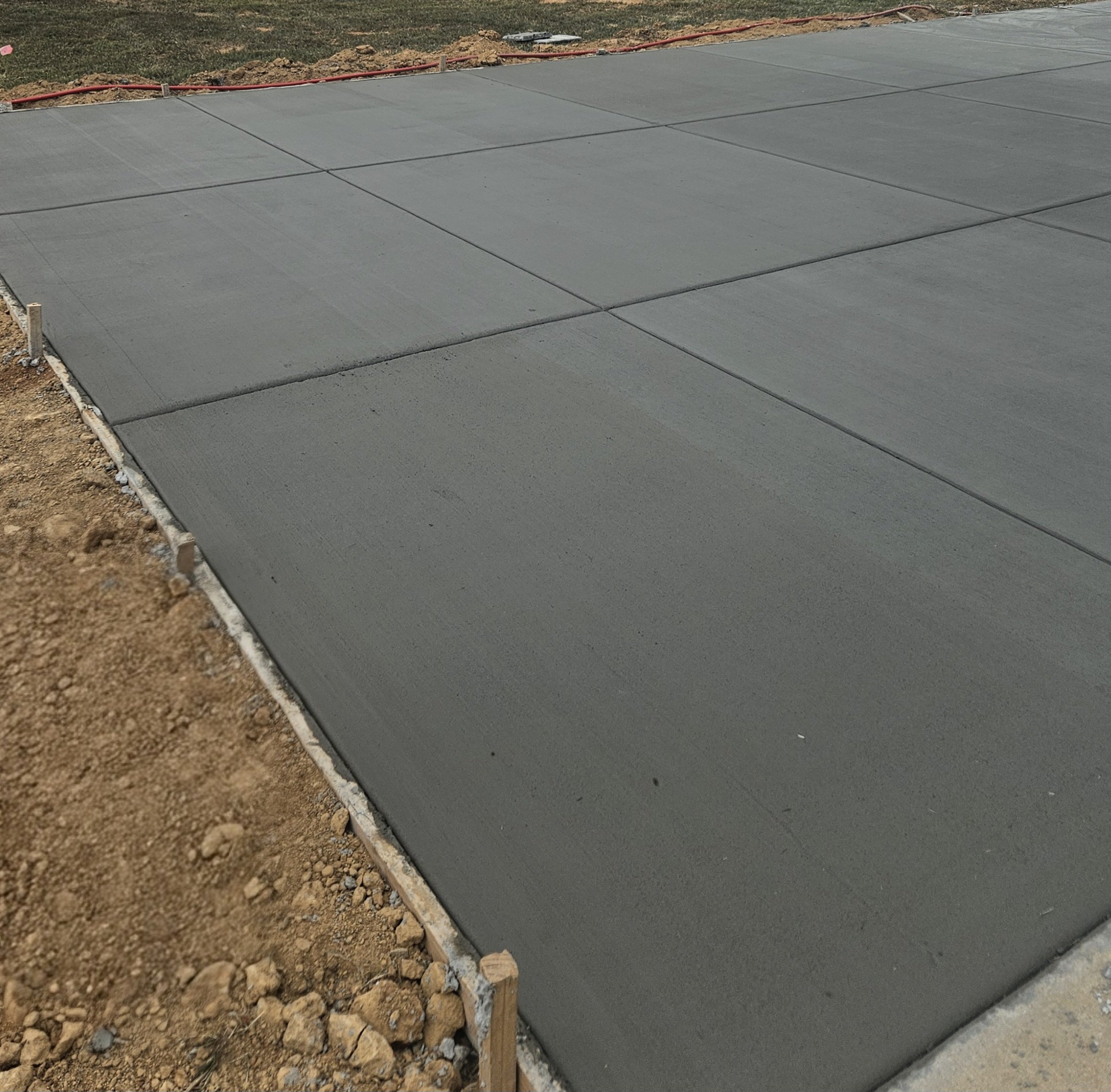 Freshly poured concrete sidewalk with wooden forms along the edges, adjacent to dirt and soil, on a construction site.