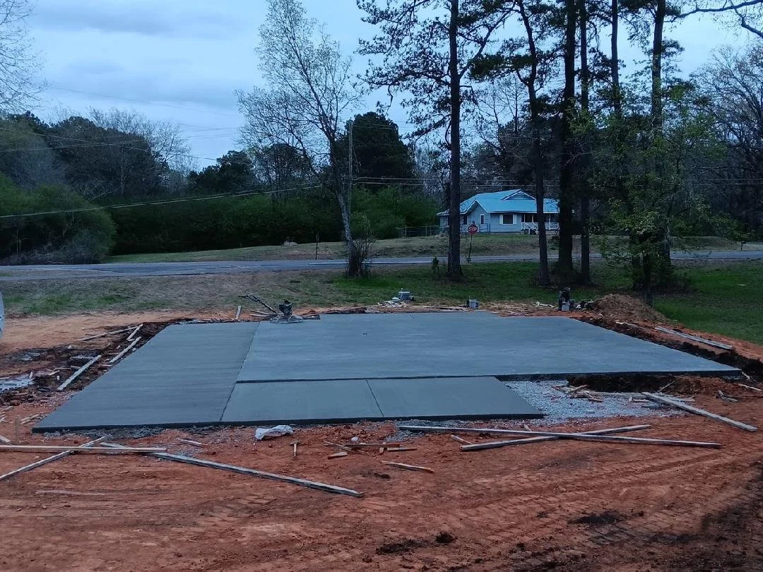 A concrete foundation is being poured for a building under construction, with some concrete slabs laid out on the site, surrounded by dirt and construction tools, with trees and a house visible in the background.