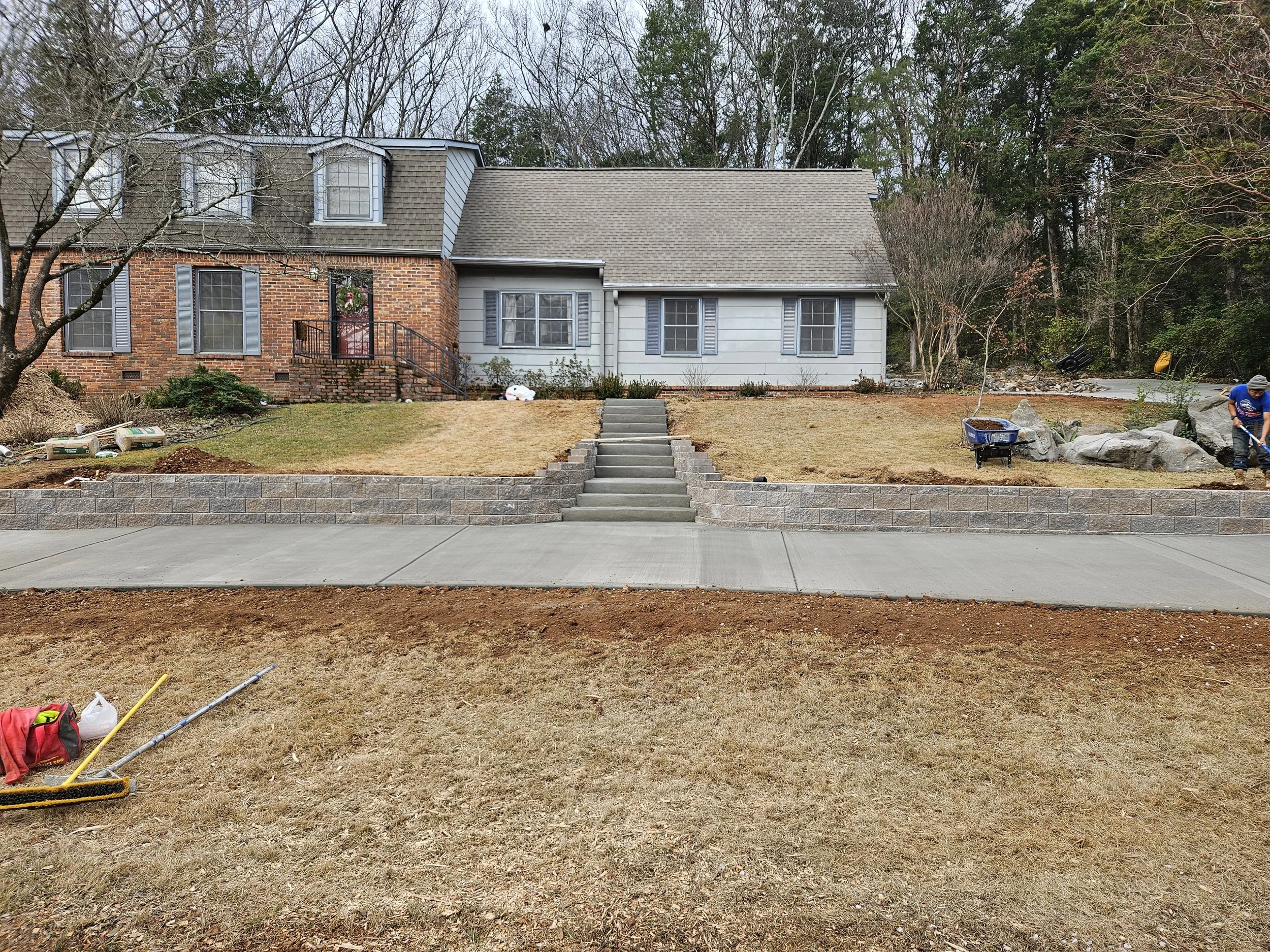 A backyard with brick and siding house, stairs leading to the house, and people working on landscaping. Items such as a wheelbarrow, tools, and bags are visible.