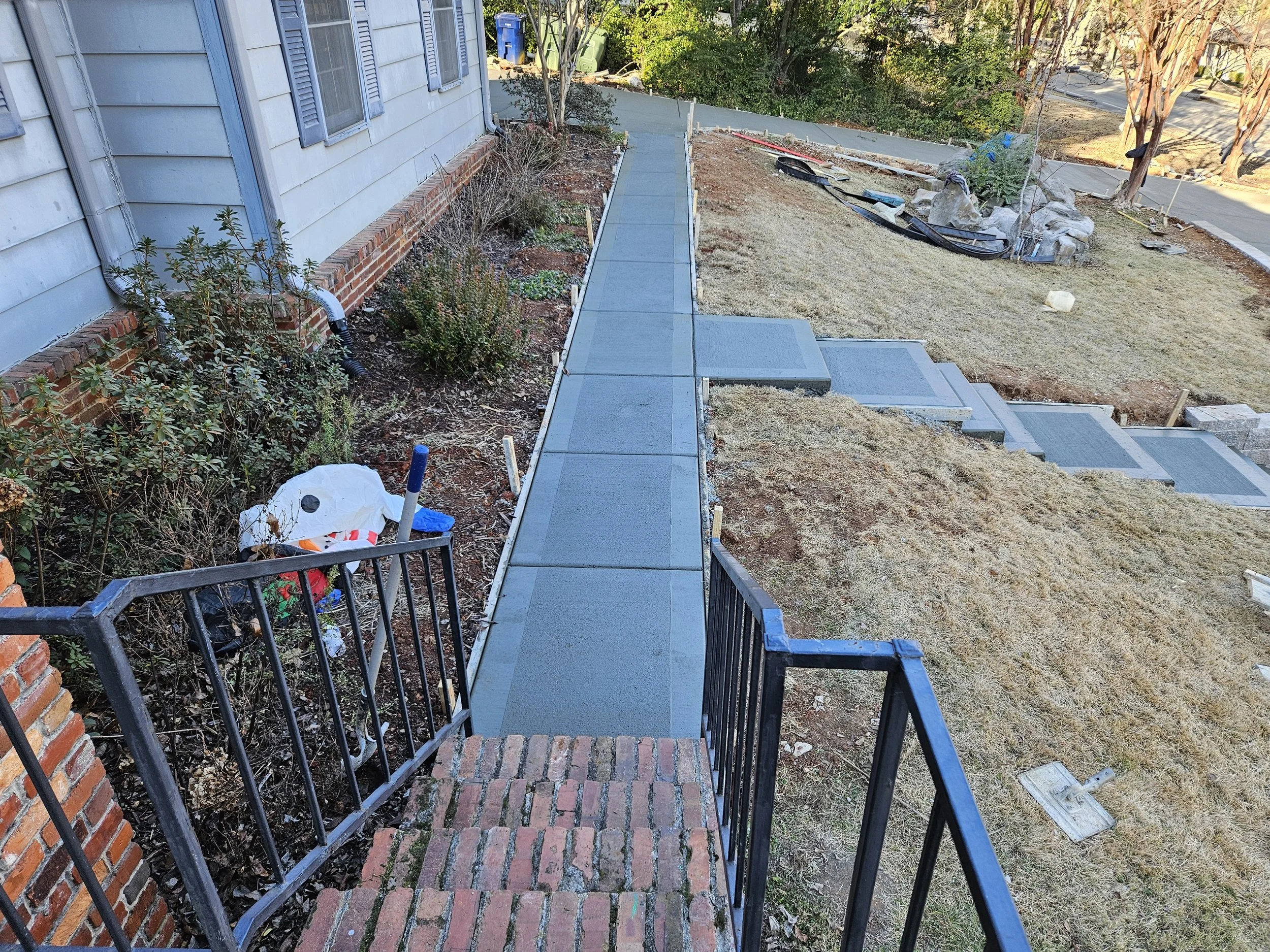 A newly poured concrete walkway with brick steps leading up to a house. There is construction equipment and dirt on the yard, and a garden area with plants and rocks.