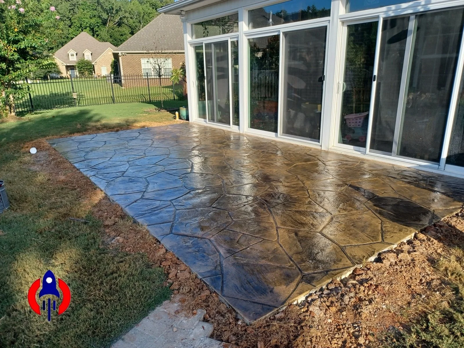A newly paved concrete patio with a stamped pattern resembling irregular stones outside a house with large glass sliding doors.