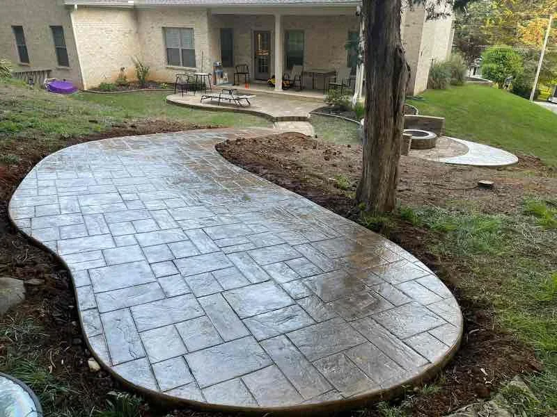 Newly constructed stamped concrete walkway in a backyard, leading to a porch with outdoor furniture, trees, and grass.