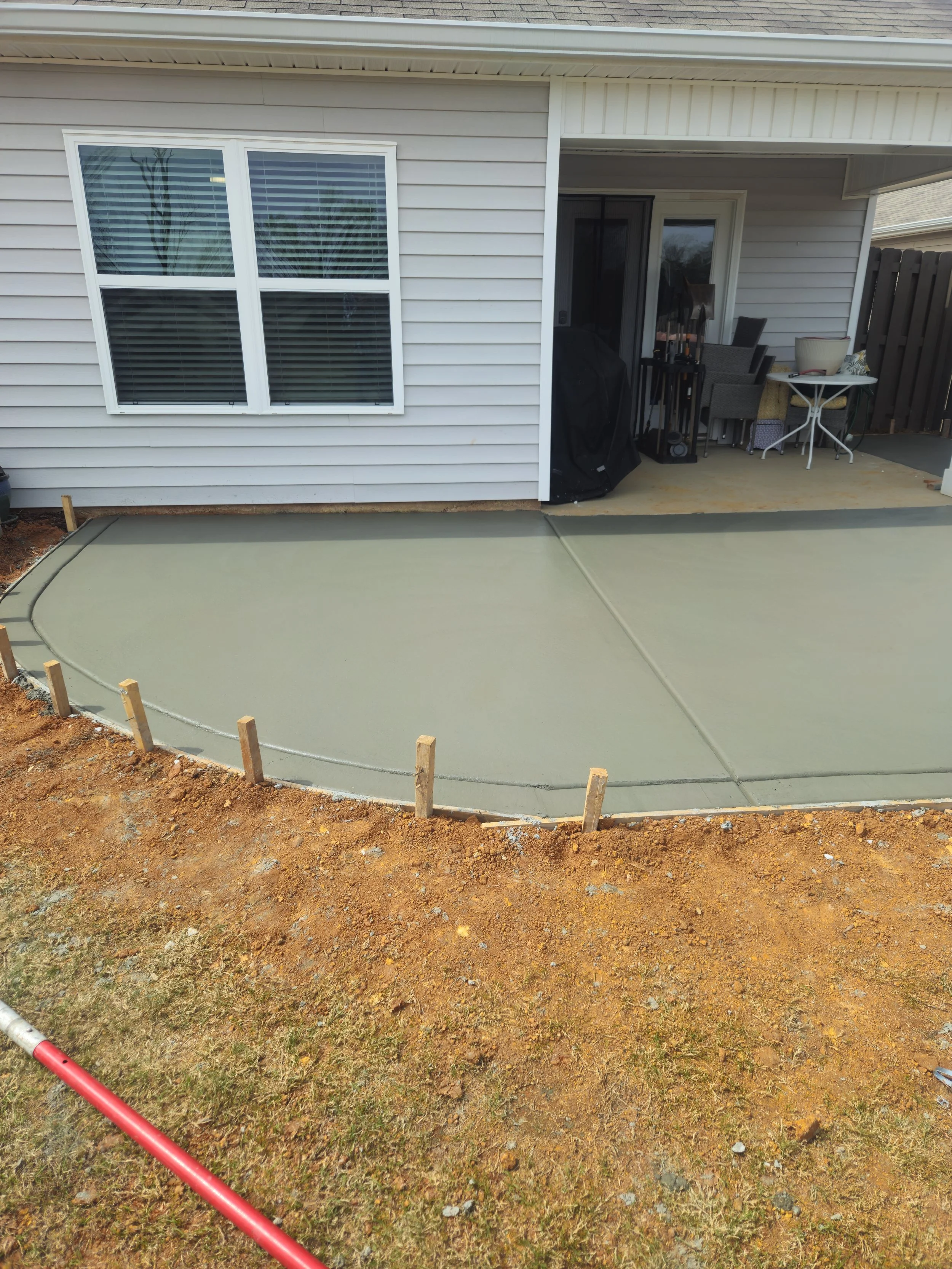 Backyard patio area with sliding glass door, patio furniture, grill covered with a black cover, and a small table with a planter. The yard has a concrete patio and a section of dirt and grass with wooden stakes outlining a new concrete slab.