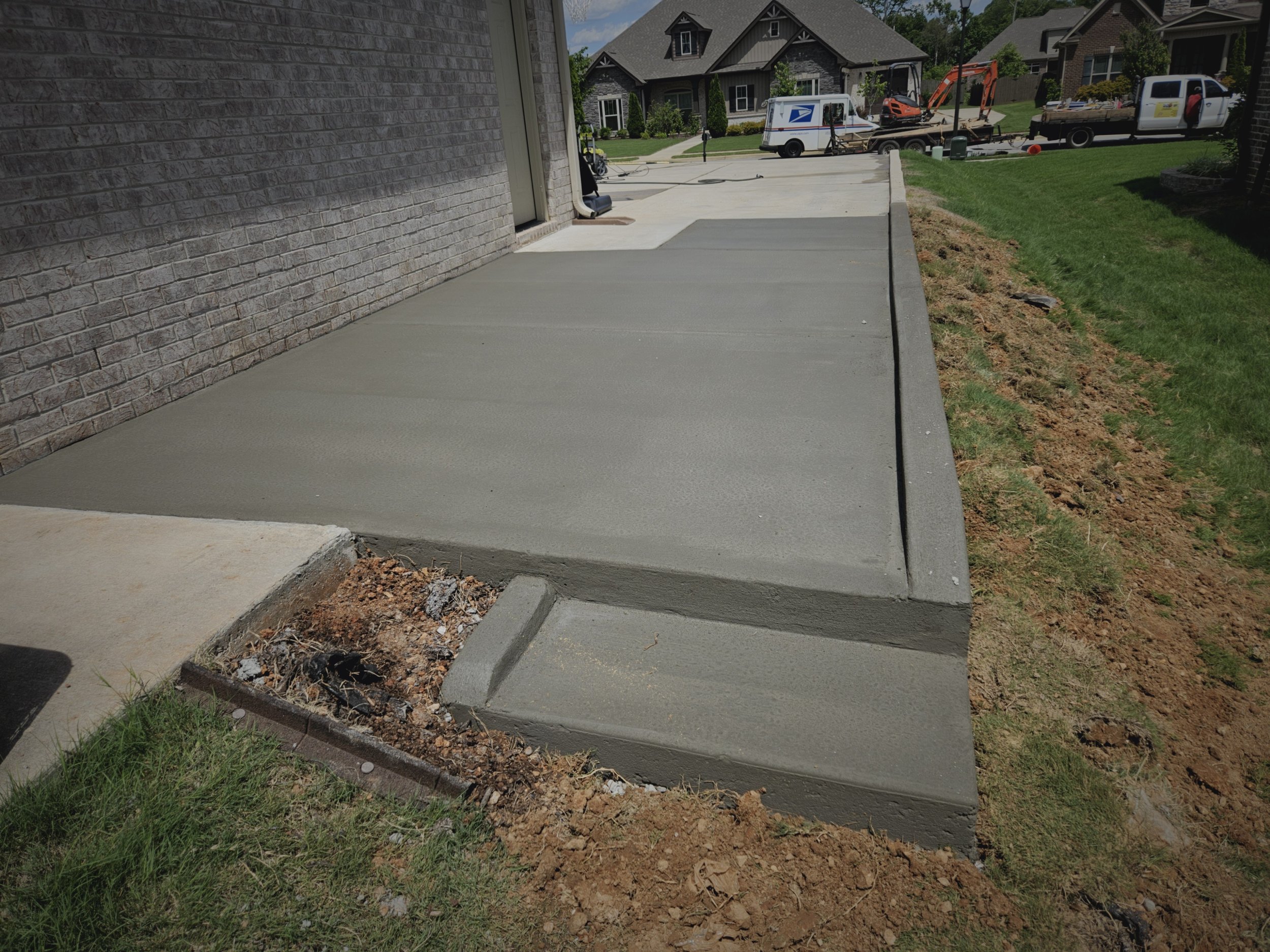 Recently poured concrete sidewalk beside a brick house with a porch, with nearby grass and dirt. In the background, there are residential houses, vehicles including a postal service truck, and a small construction excavator.