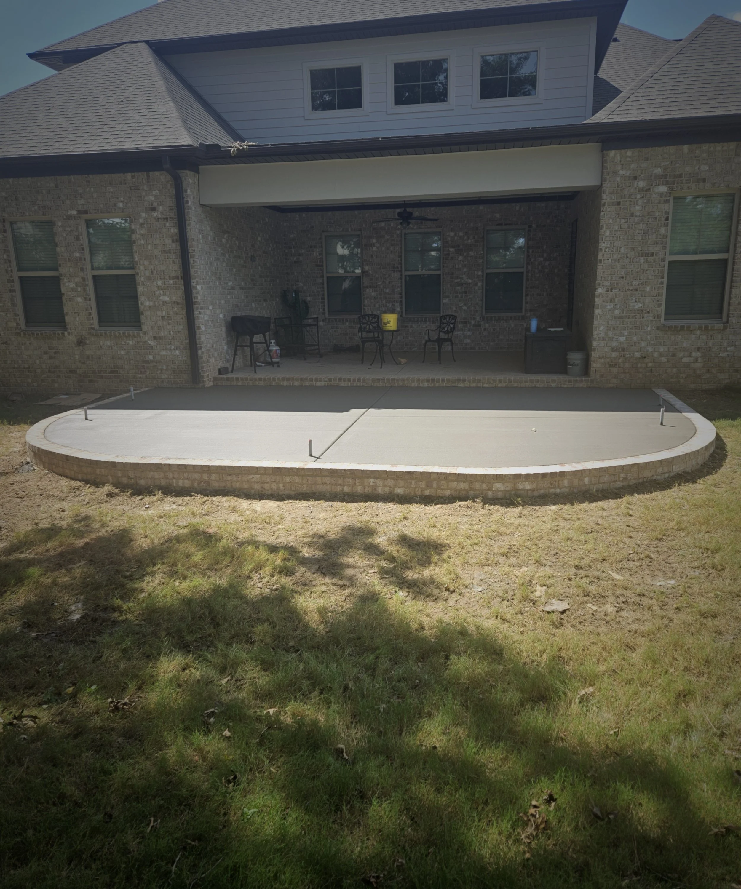 Backyard patio with a concrete slab, brick wall house with three windows, a yellow bucket, black chairs, a black table, and some outdoor items.
