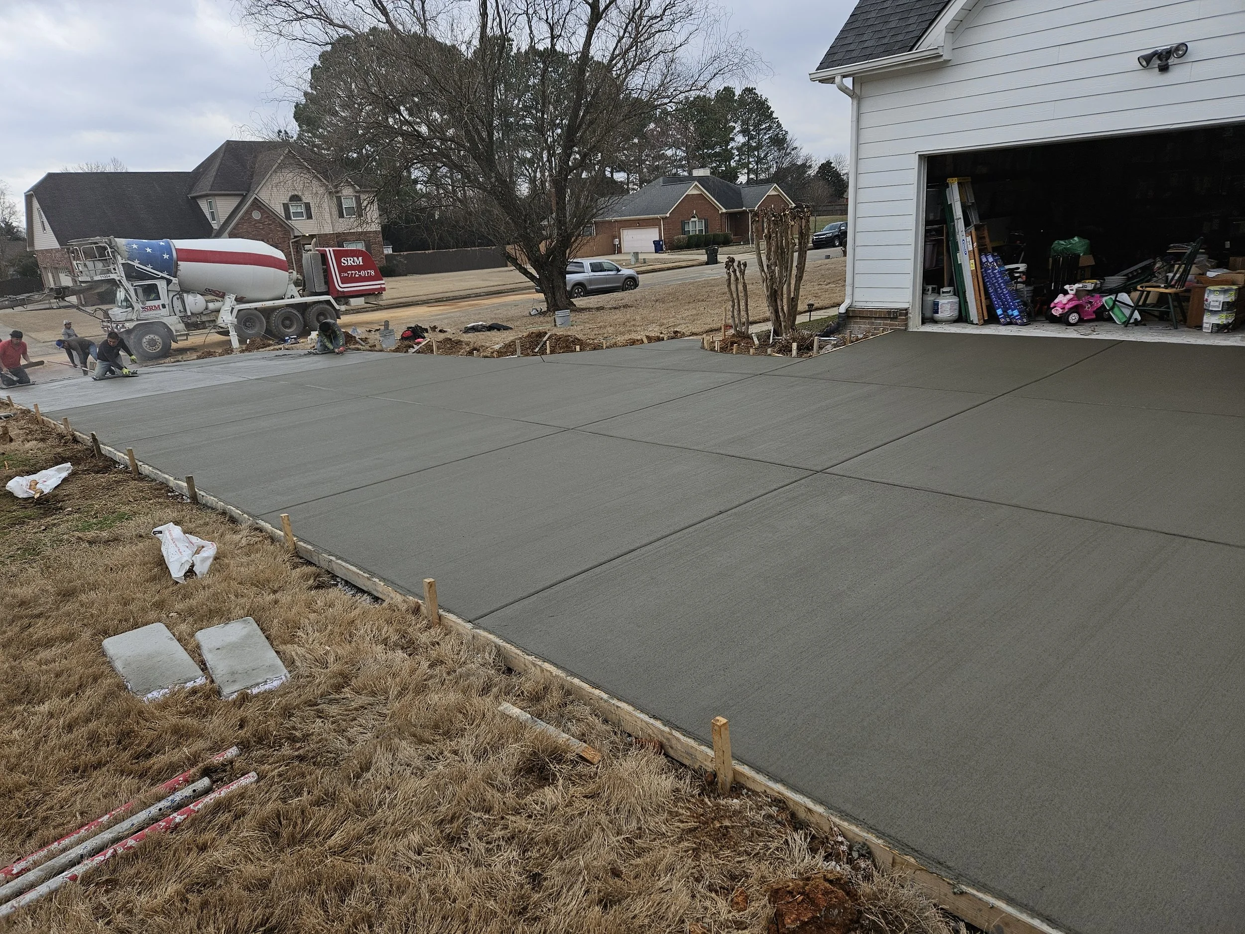 Workers are pouring and smoothing concrete for a driveway in a residential neighborhood. A cement mixer truck is present, and the scene shows a garage on the right and houses across the street.