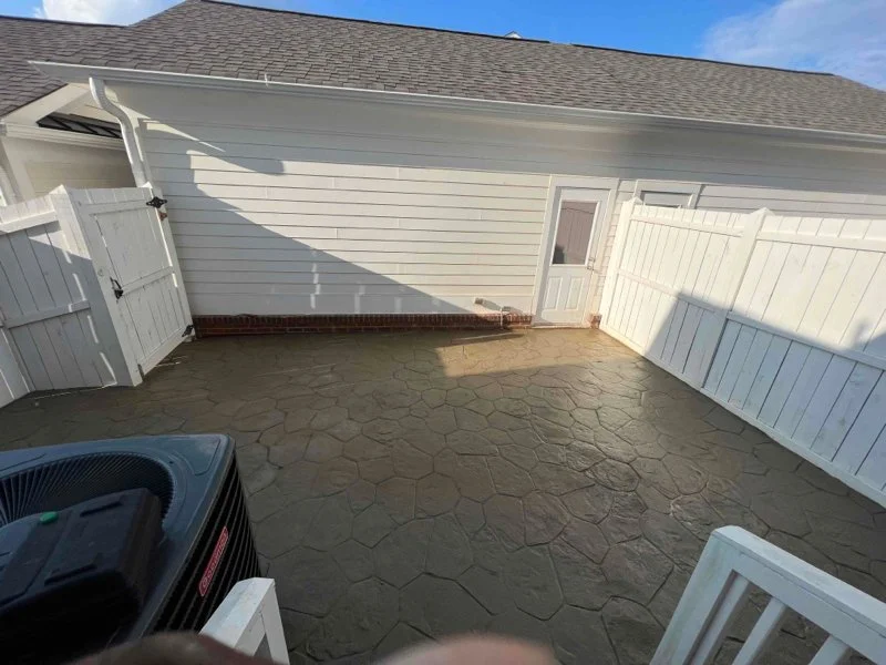 Empty backyard patio with stamped concrete, white wooden fence, small door, and house wall in background under blue sky.