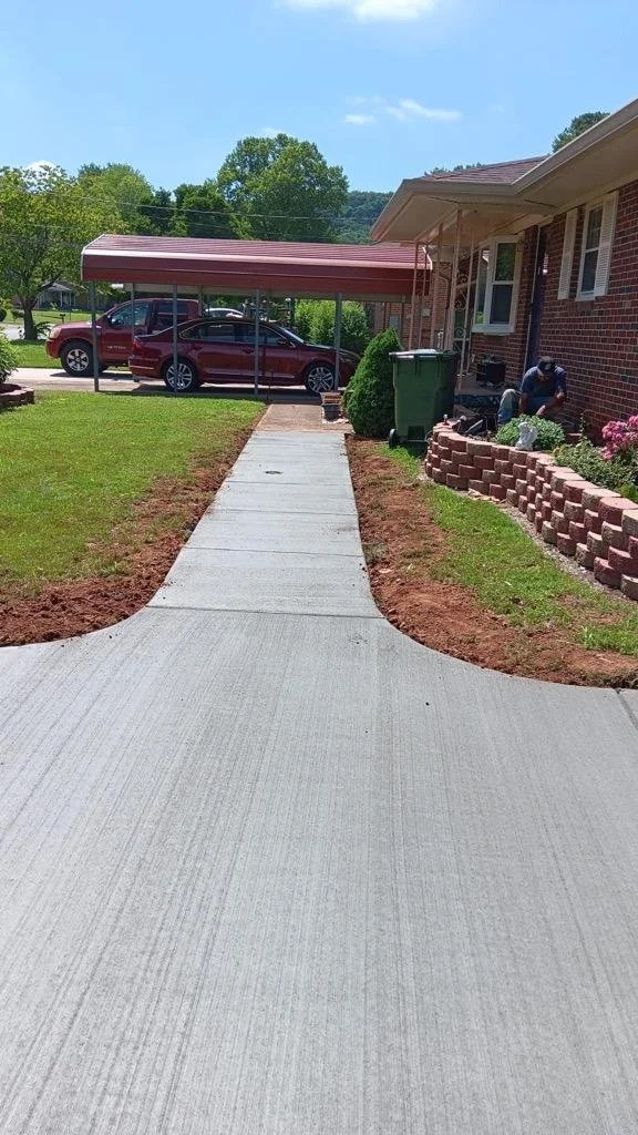 Concrete sidewalk leading from the street to the porch of a brick house with a carport and a parked maroon vehicle. A man is working near the house with gardening tools, and there are bushes, a flower bed, and a green trash can beside the house.