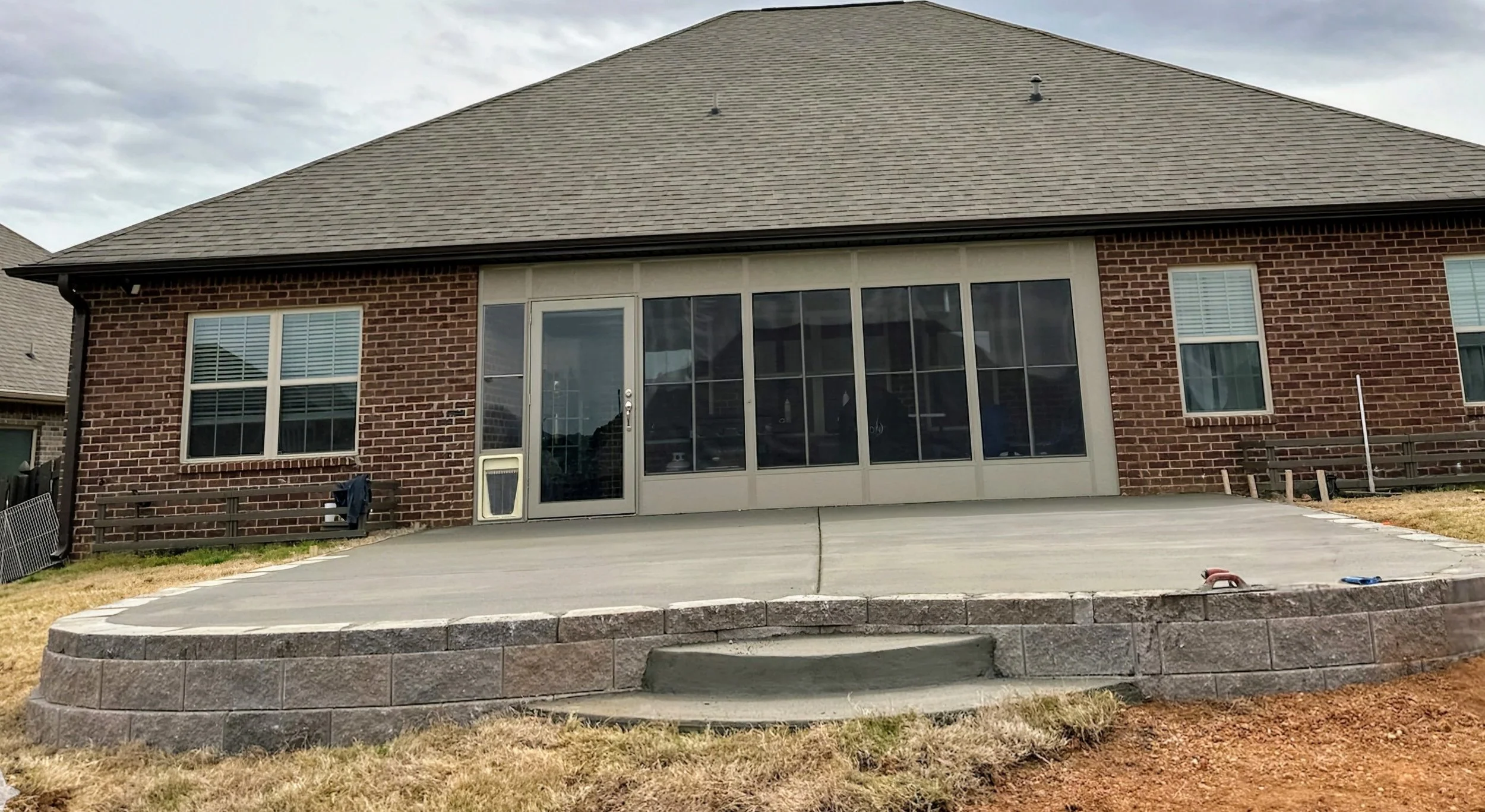Back of a brick house with large screened porch, two windows, and a gray shingle roof. There is a concrete patio and a curved stone retaining wall in the yard.