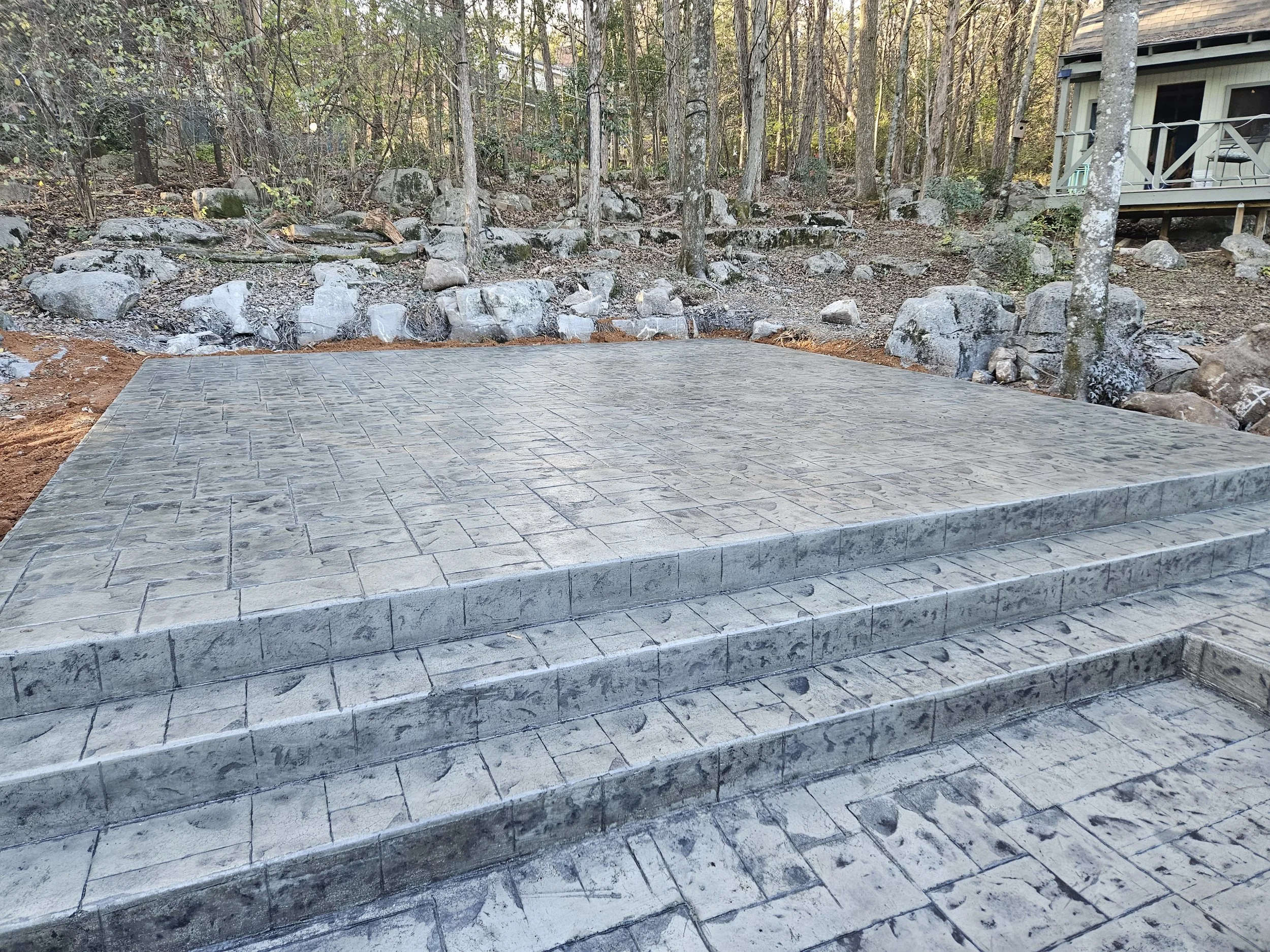 Newly constructed stone patio with steps leading up into a wooded backyard, featuring trees, rocks, and part of a house with a deck.