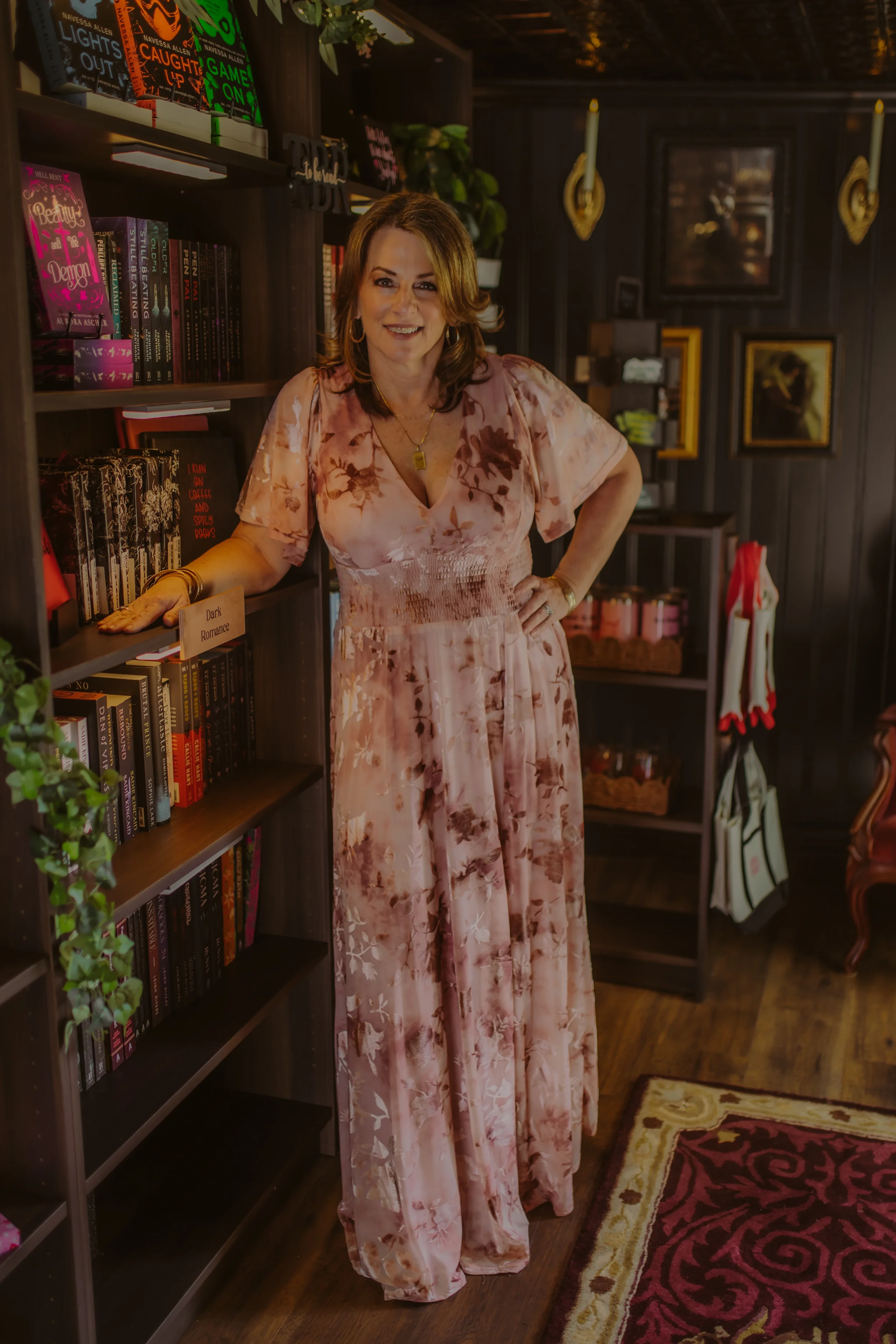 A woman in a pink floral maxi dress smiling inside a cozy library or bookstore with dark wood shelves filled with books, framed pictures, and decorative candles hanging on the wall.