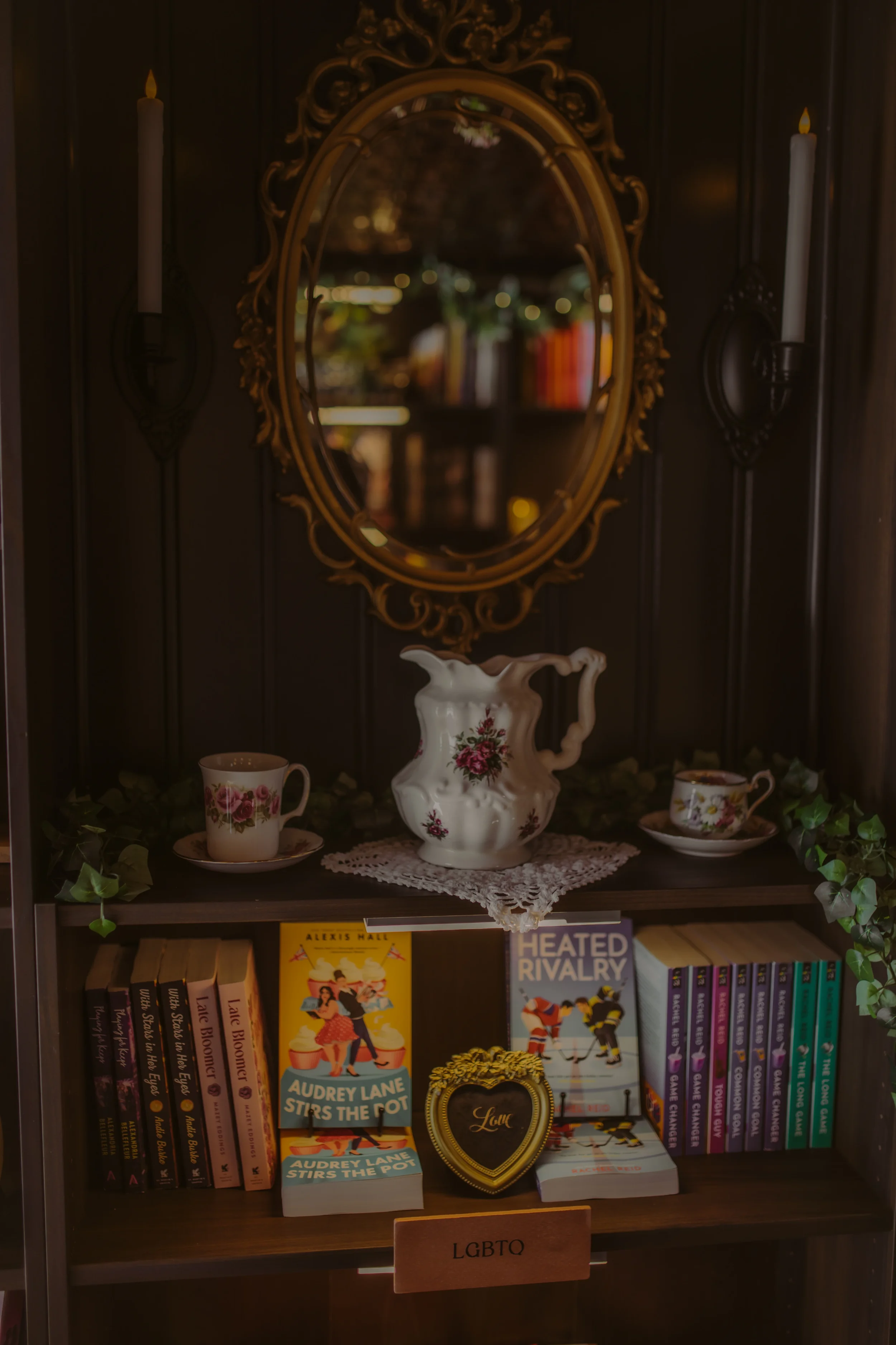 A bookshelf with several books, a decorative heart-shaped item, and a sign labeled 'LGBTQ'. The top of the bookshelf features a mirror with an ornate gold frame, a porcelain pitcher with a floral design, and two coffee cups with matching floral patterns. The background includes a mirror reflecting books and furnishings.