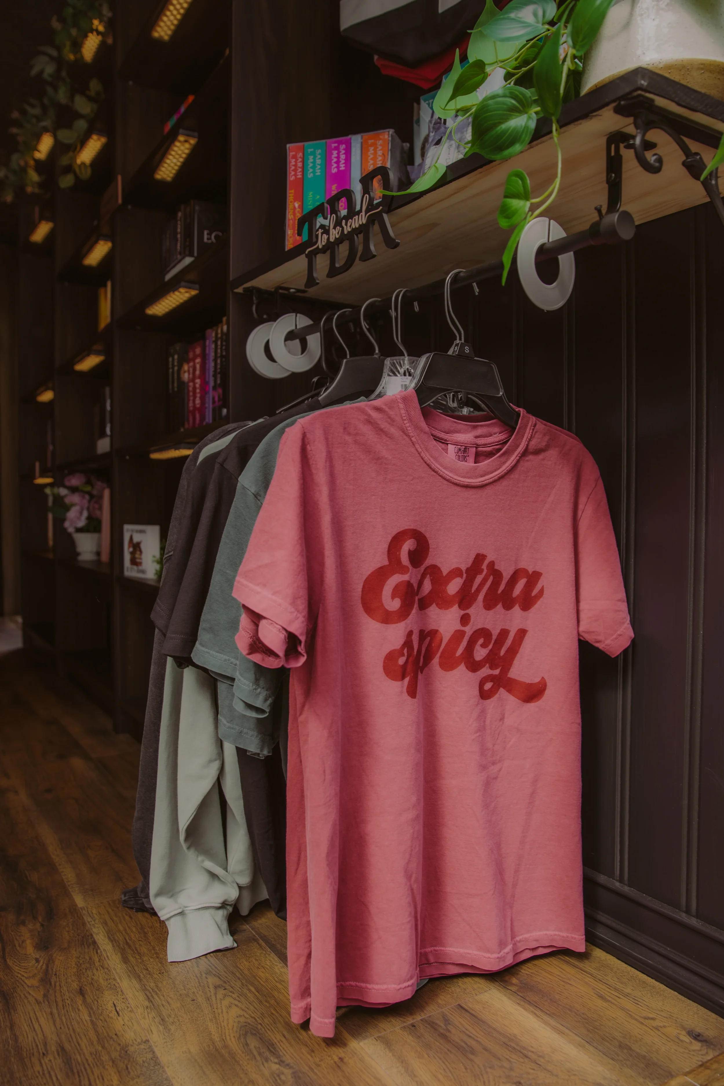 A clothing rack displaying a pink t-shirt with the word 'Extra Spicy' printed on it, hanging among other shirts in a store with wooden flooring and dark shelving.