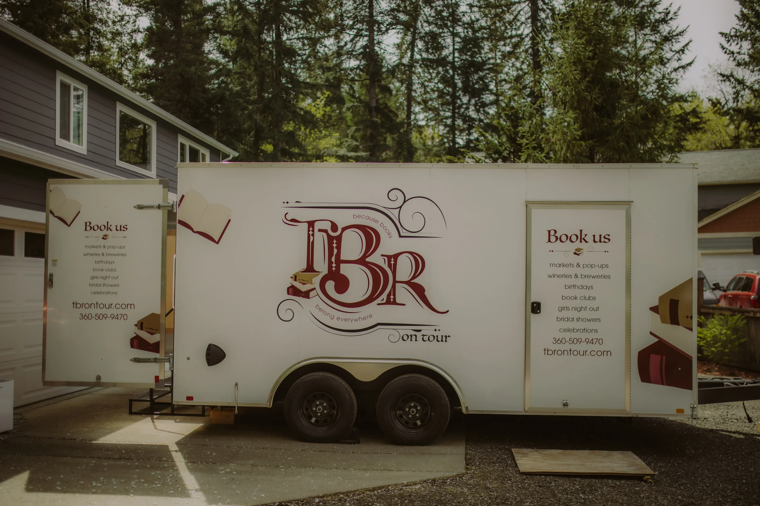 A white book-themed mobile trailer parked on a driveway with signs displaying various event services, with houses and tall trees in the background.