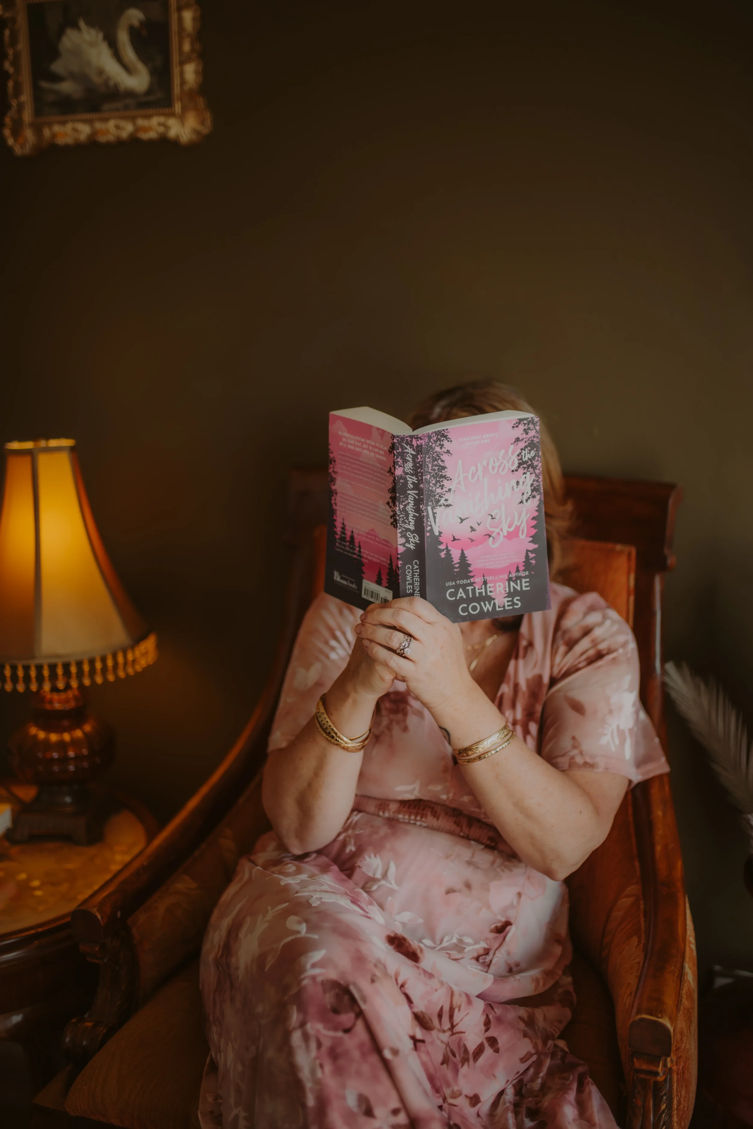 A woman wearing a pink floral dress is sitting on a wooden chair with a carved backrest, reading a book titled 'Across the Wild Sky'. She is accessorized with gold jewelry including bracelets and rings. The background features a brown wall, a framed artwork of a swan, and a wooden nightstand with a yellow lampshade and some small objects. The setting has warm ambient lighting.