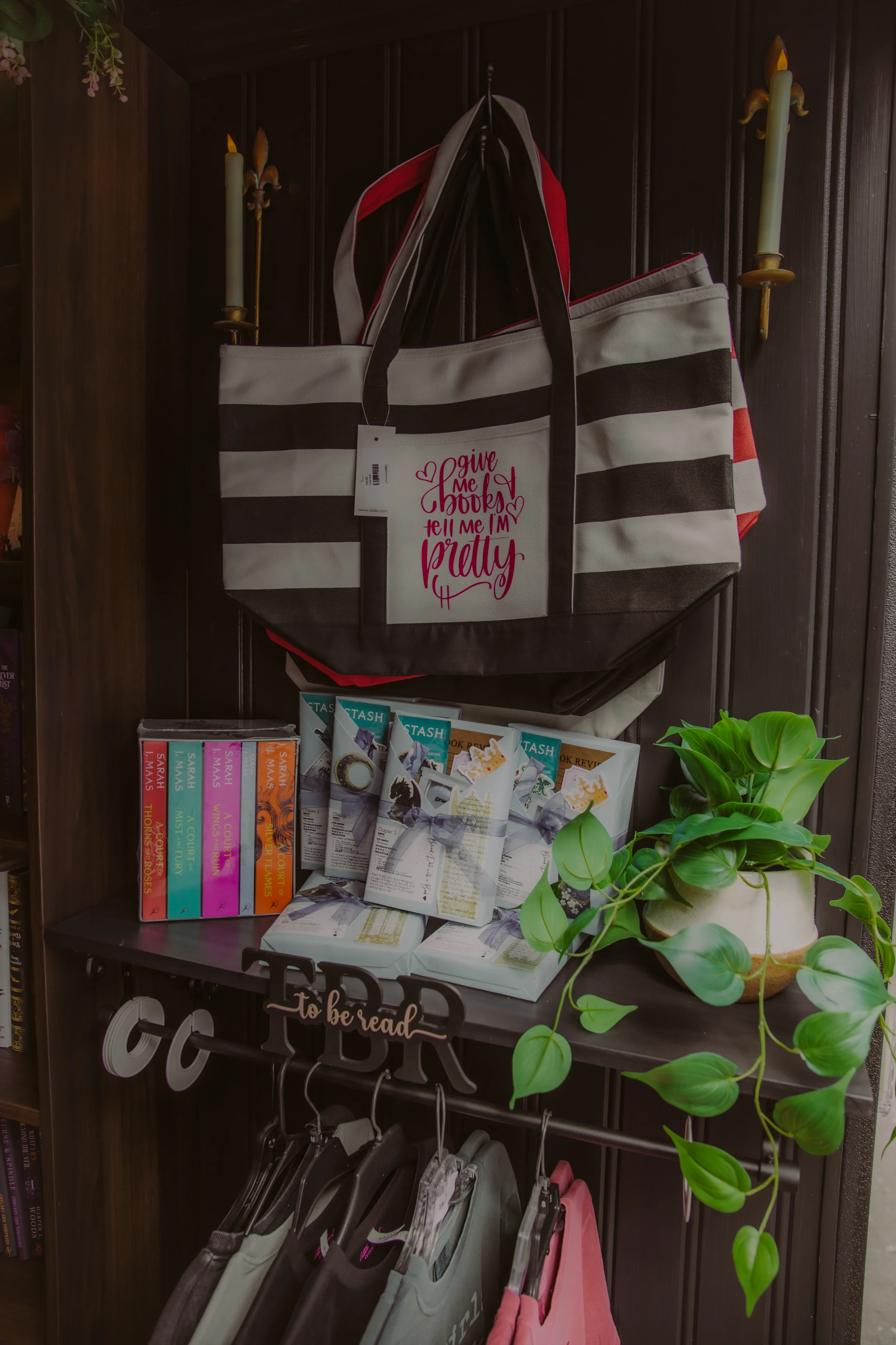 A tote bag with black and white horizontal stripes hanging on a hook, with a tag that says "give me books, tell me I'm pretty." Behind the tote bag, there are two-folded bags with red and black colors. To the left, a collection of books is visible, and a lush green plant with rounded leaves is in the bottom right corner. The background features dark wooden paneling with wall-mounted candle holders.