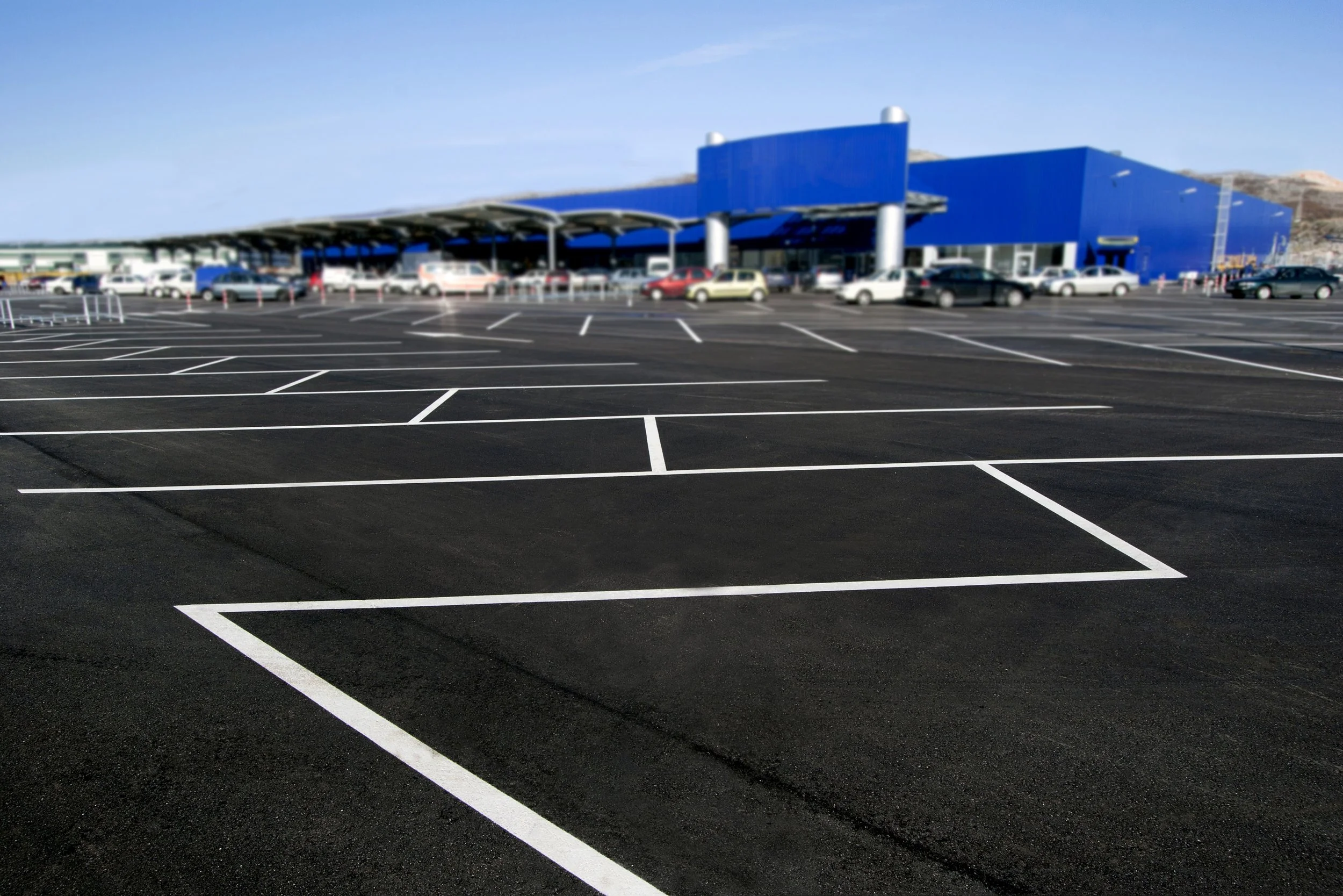 Newly painted parking lot in front of a large store with a blue exterior and a parking structure, cars parked along the building and in the lot.