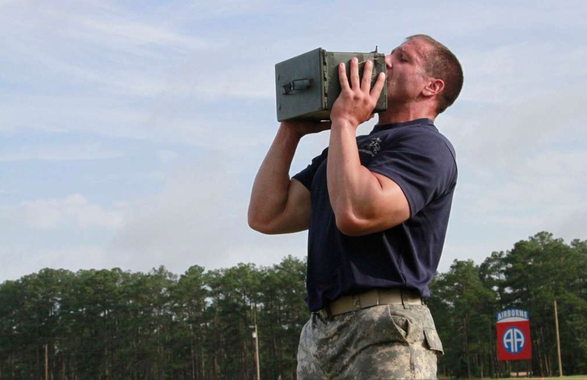 As a soldier, Mike is dressed in camouflage pants and a navy blue T-shirt is aiming a handheld radio or communications device towards the sky during daytime with cloudy skies in the background.
