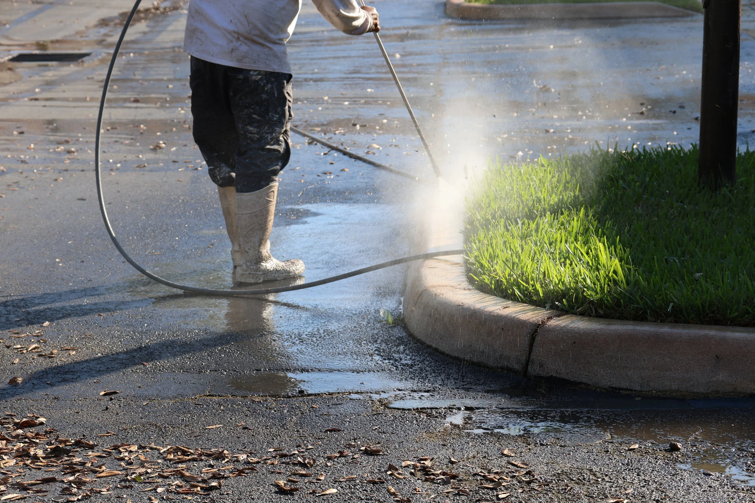 Pressure washing a sidewalk near a grassy area, wearing rubber boots and work clothes, with water spray visible.