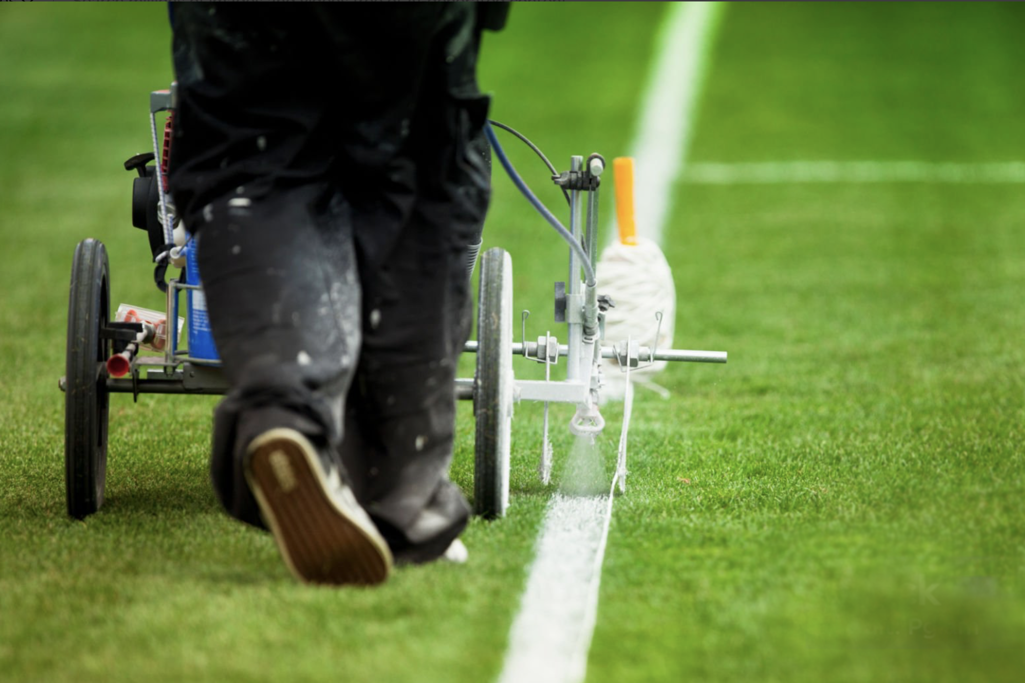 Person operating a line marking machine on a green field, applying white paint to mark lines.