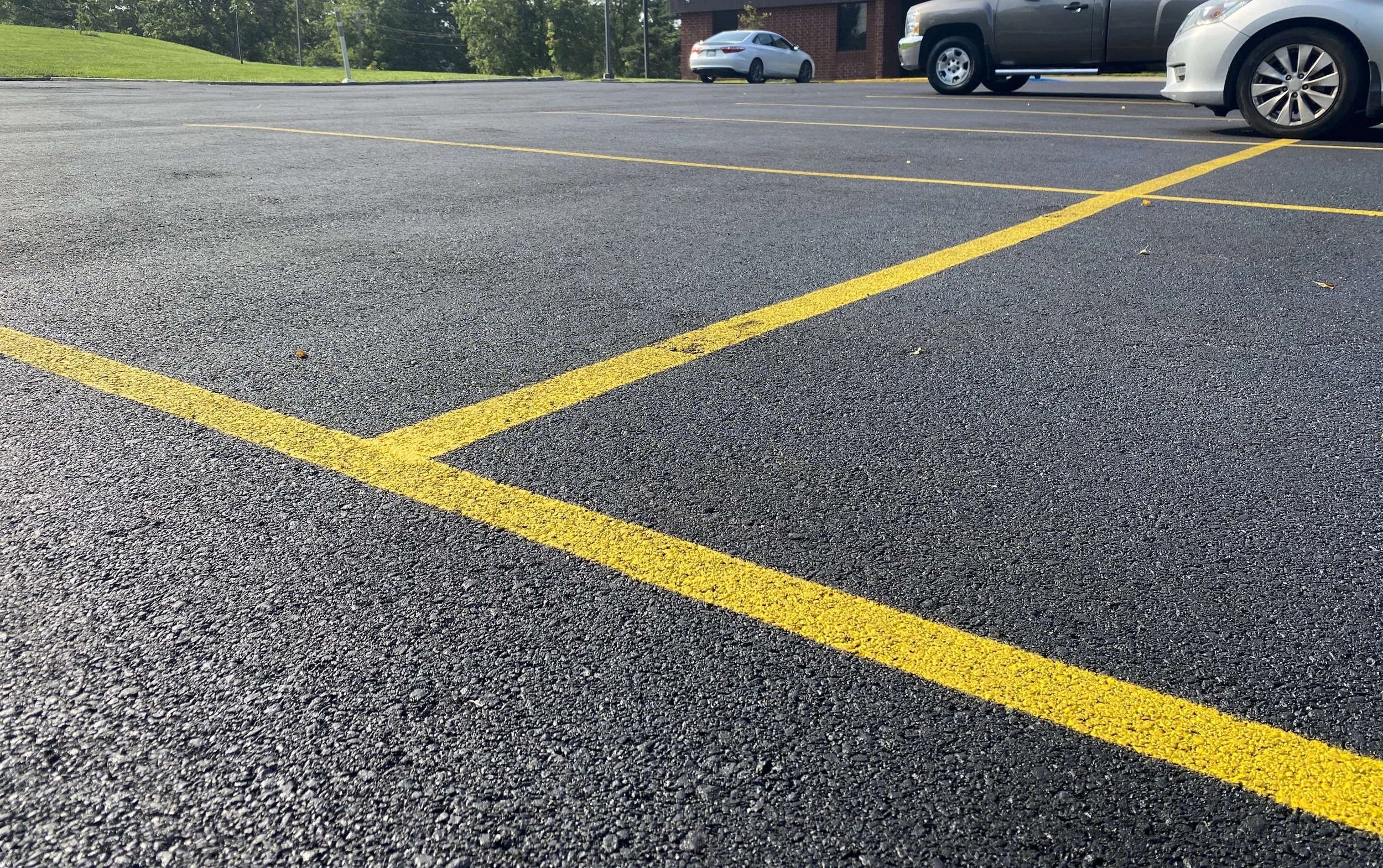 Parking lot with yellow painted lines marking parking spaces, a few parked cars, a grassy hill, and a brick building in the background.