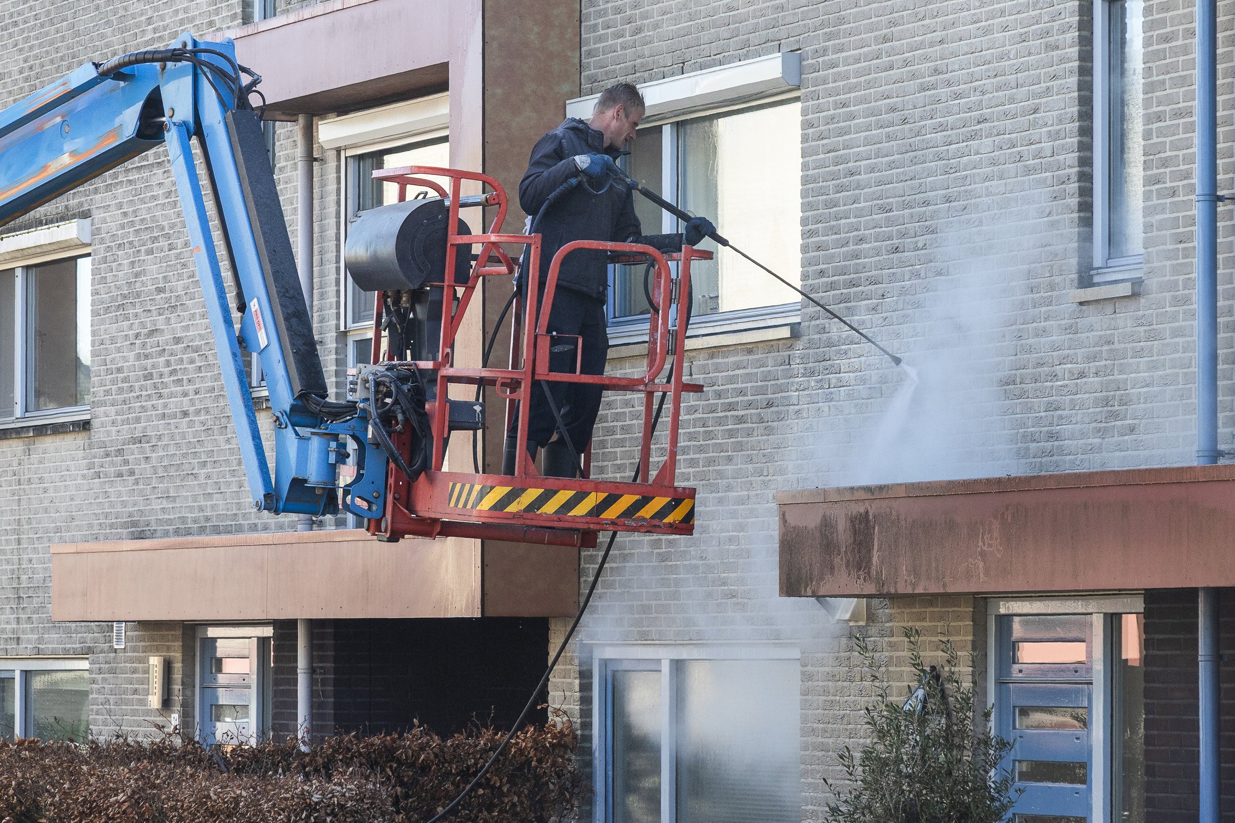 A man in black work clothes cleaning the exterior windows of a multi-story brick apartment building using a pressure washer.