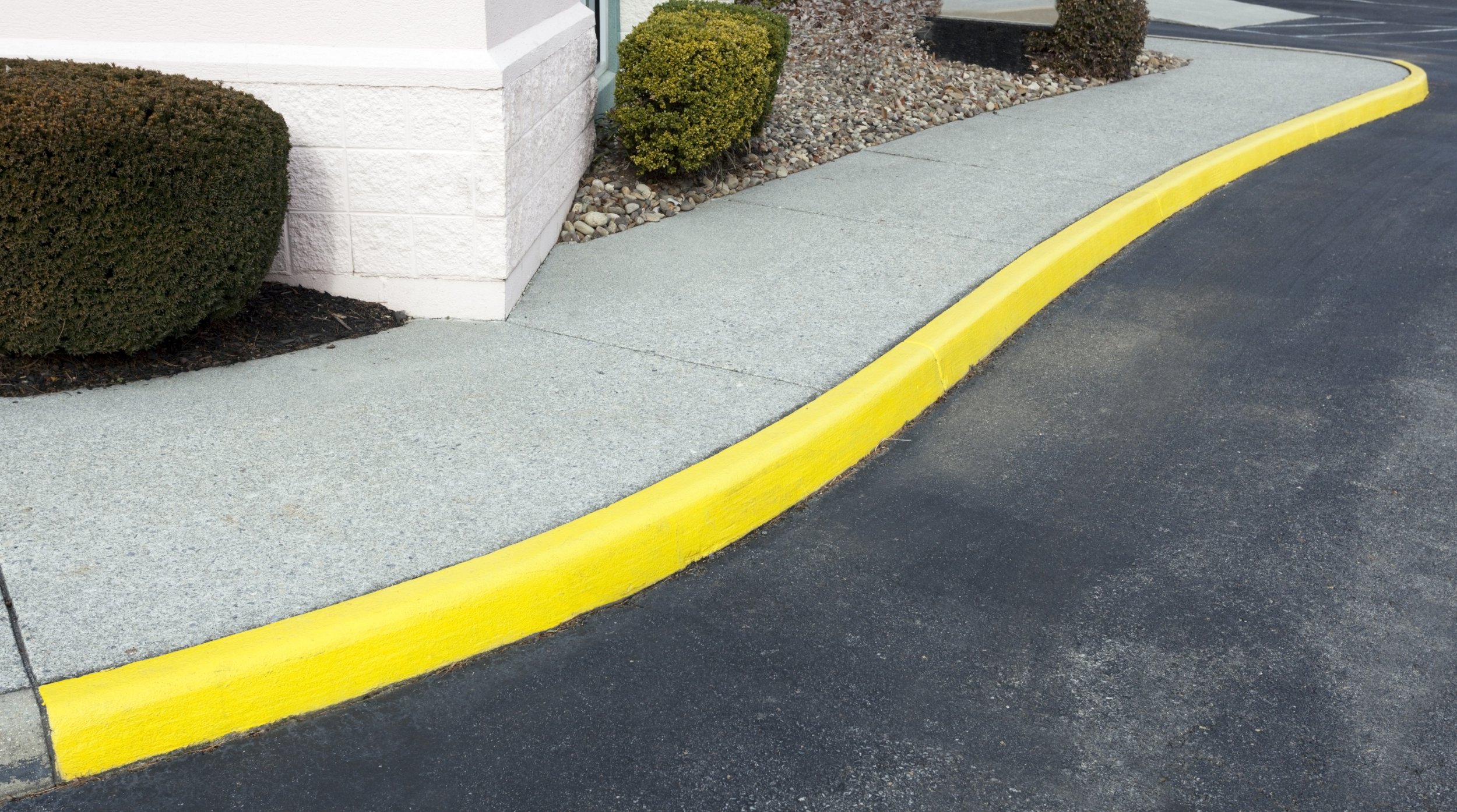 Concrete sidewalk with a yellow curb next to a parking lot, with bushes and gravel near a building wall.
