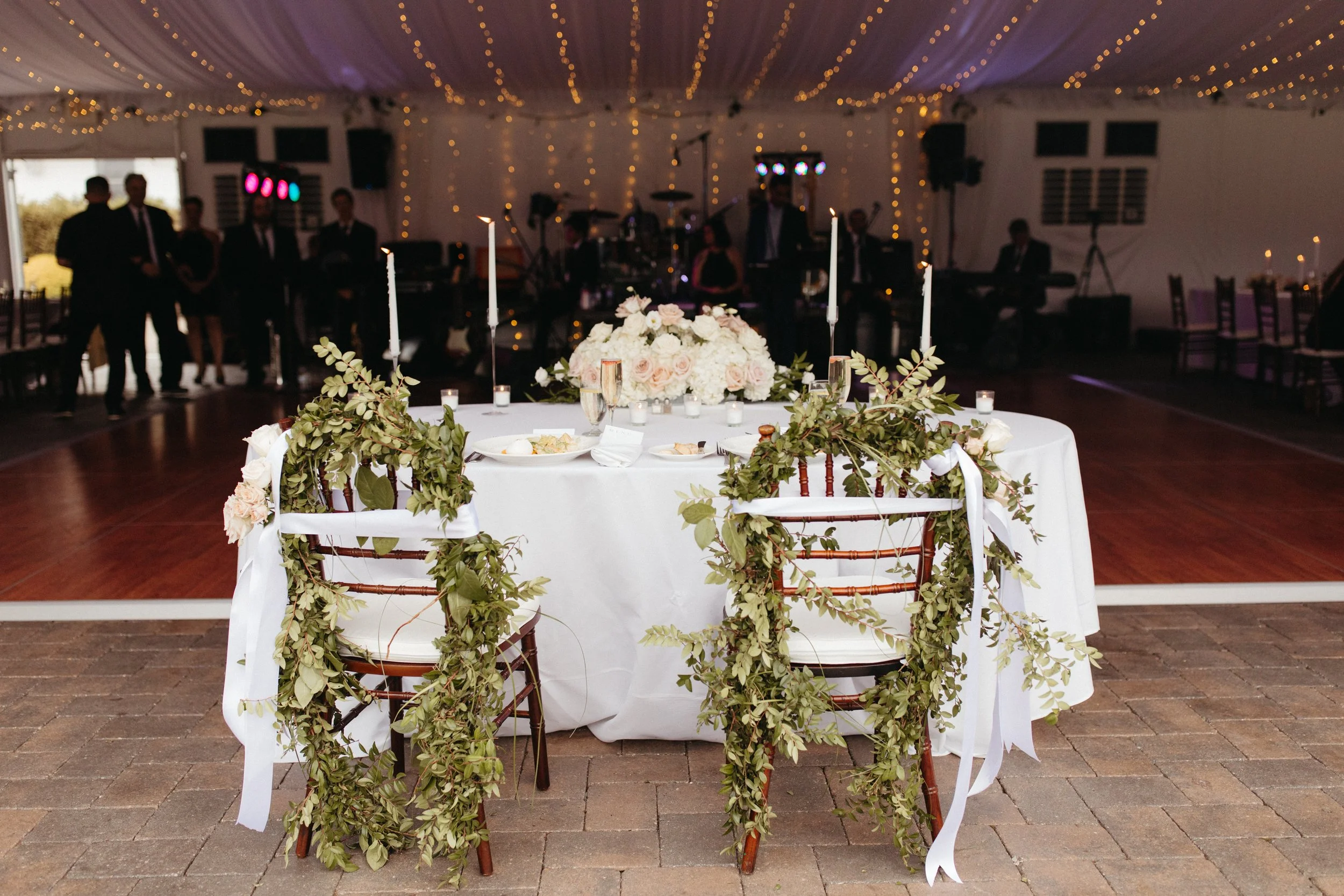 A decorated wedding reception table with floral arrangements and greenery on white tablecloth, surrounded by chairs, with a band and string lights in the background during the evening.