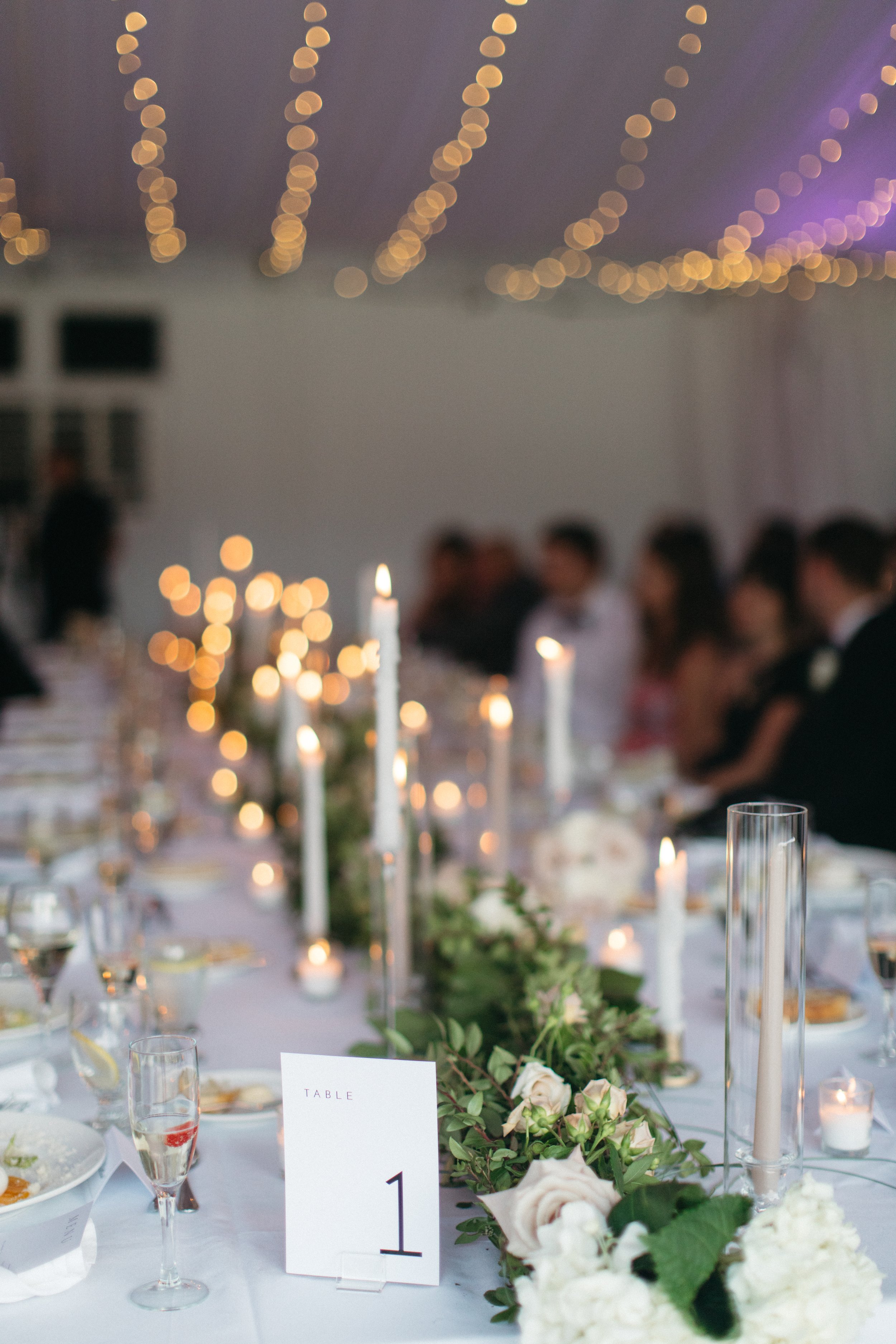 A wedding reception table decorated with candles, white flowers, and greenery, with guests seated in the background under warm string lighting, creating an elegant ambiance.