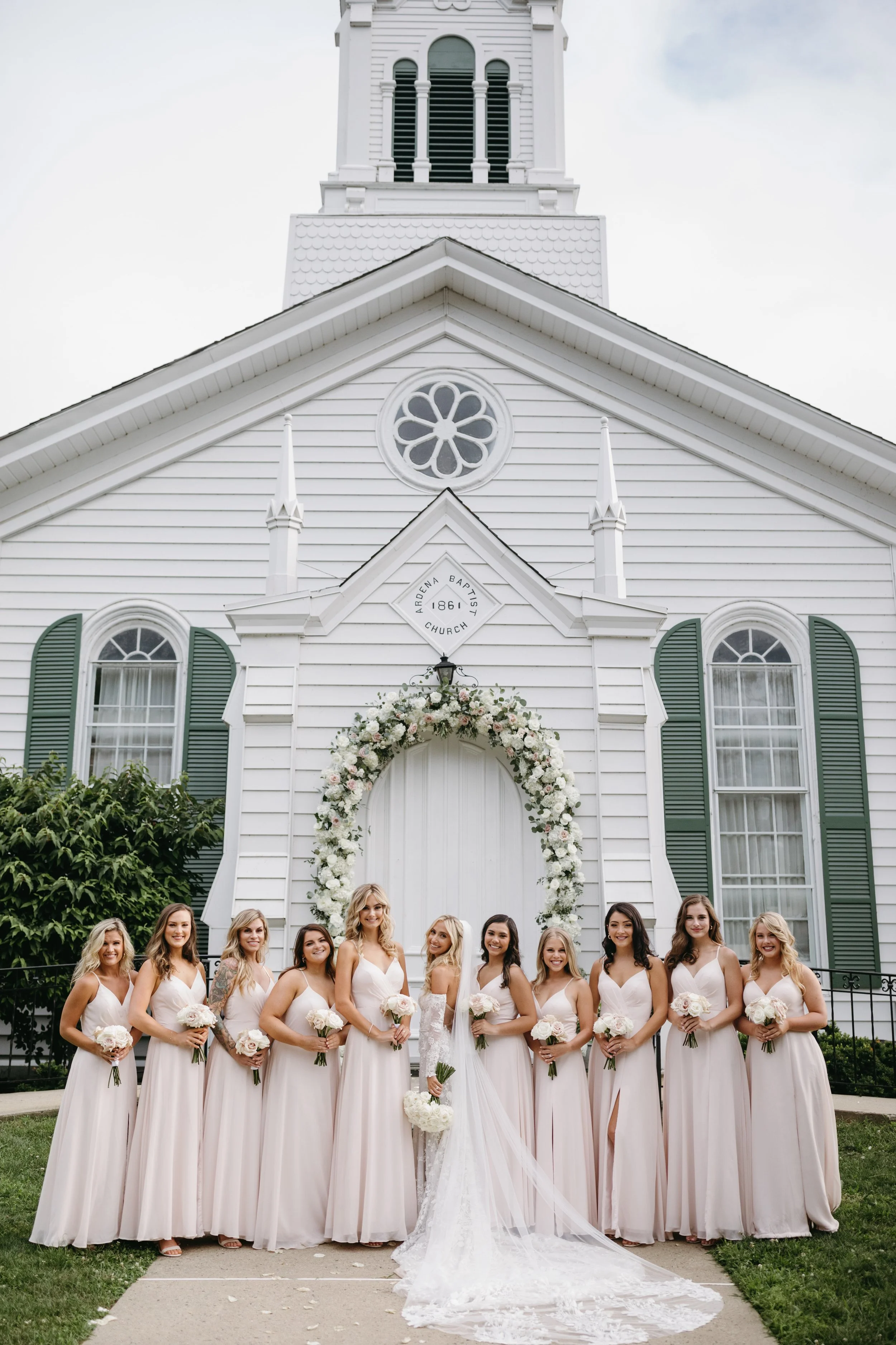 Bride and bridesmaids standing in front of a white church with floral wreath, holding bouquets of white flowers, dressed in matching light pink dresses, on a cloudy day.