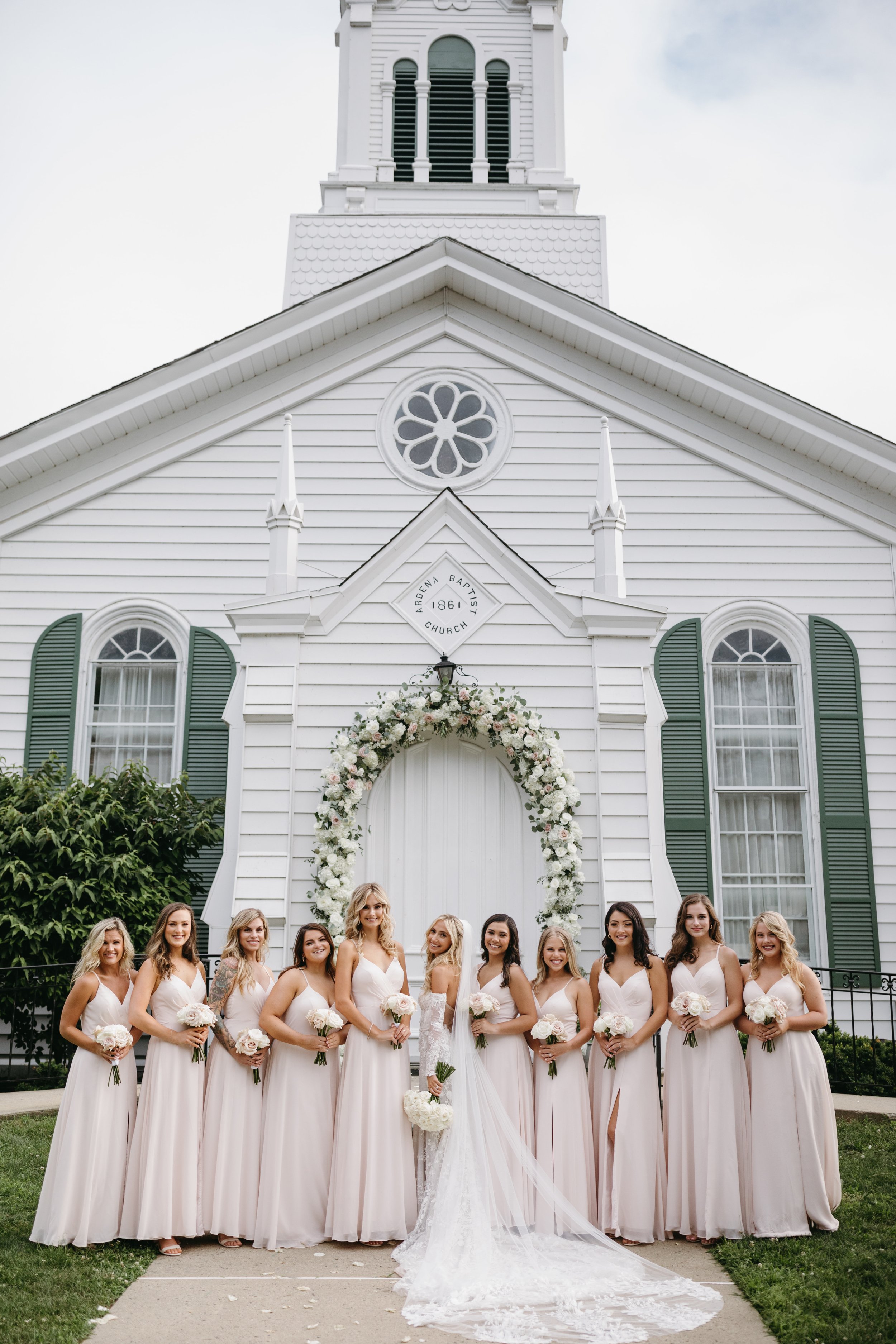 A group of women in white dresses are standing in front of a white church with floral decorations, likely for a wedding.