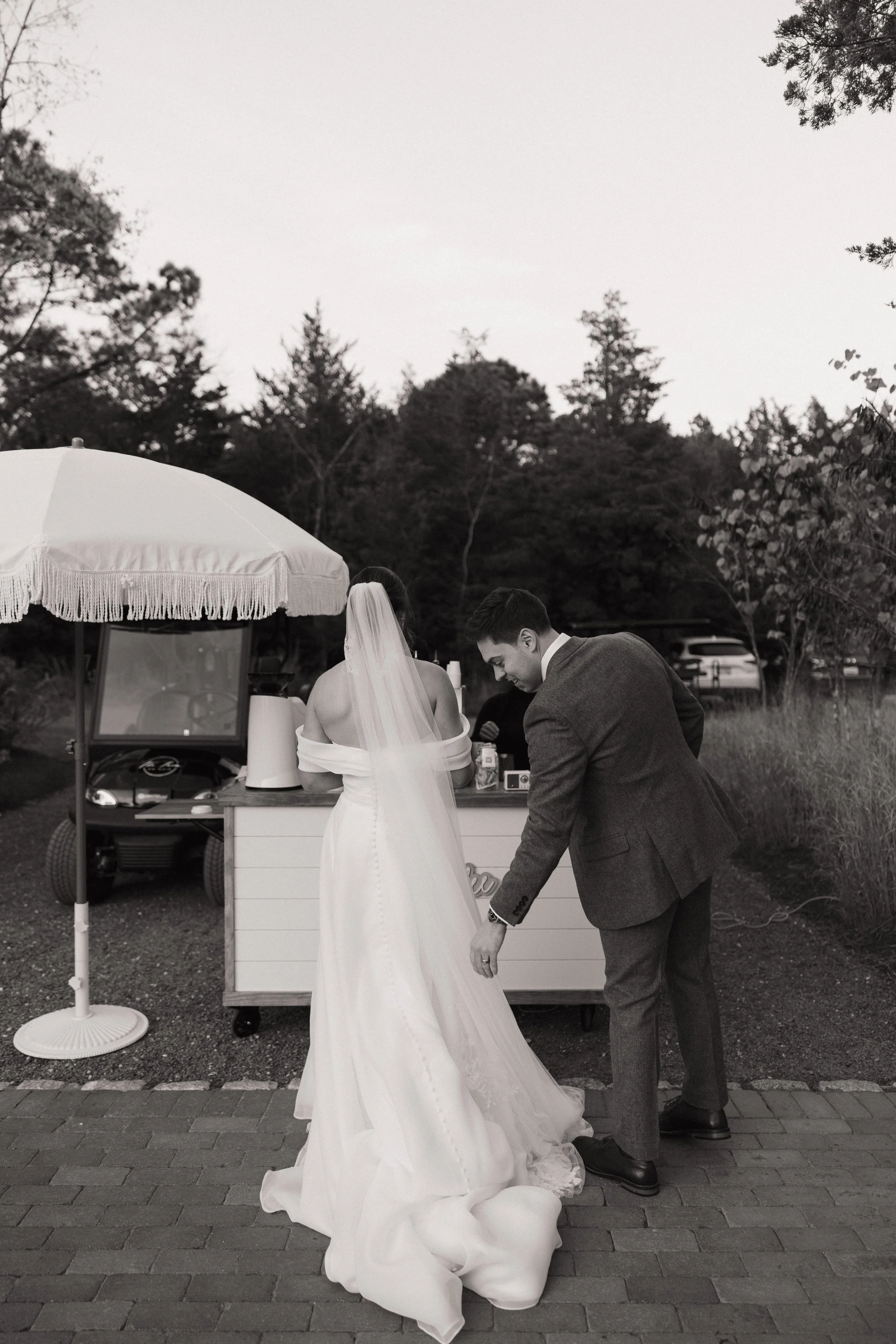 A bride in a wedding dress and veil stands next to a groom in a suit, both leaning over a small wooden cart or table outdoors, with a large umbrella overhead. They are next to a wooded area and a golf cart is visible in the background.