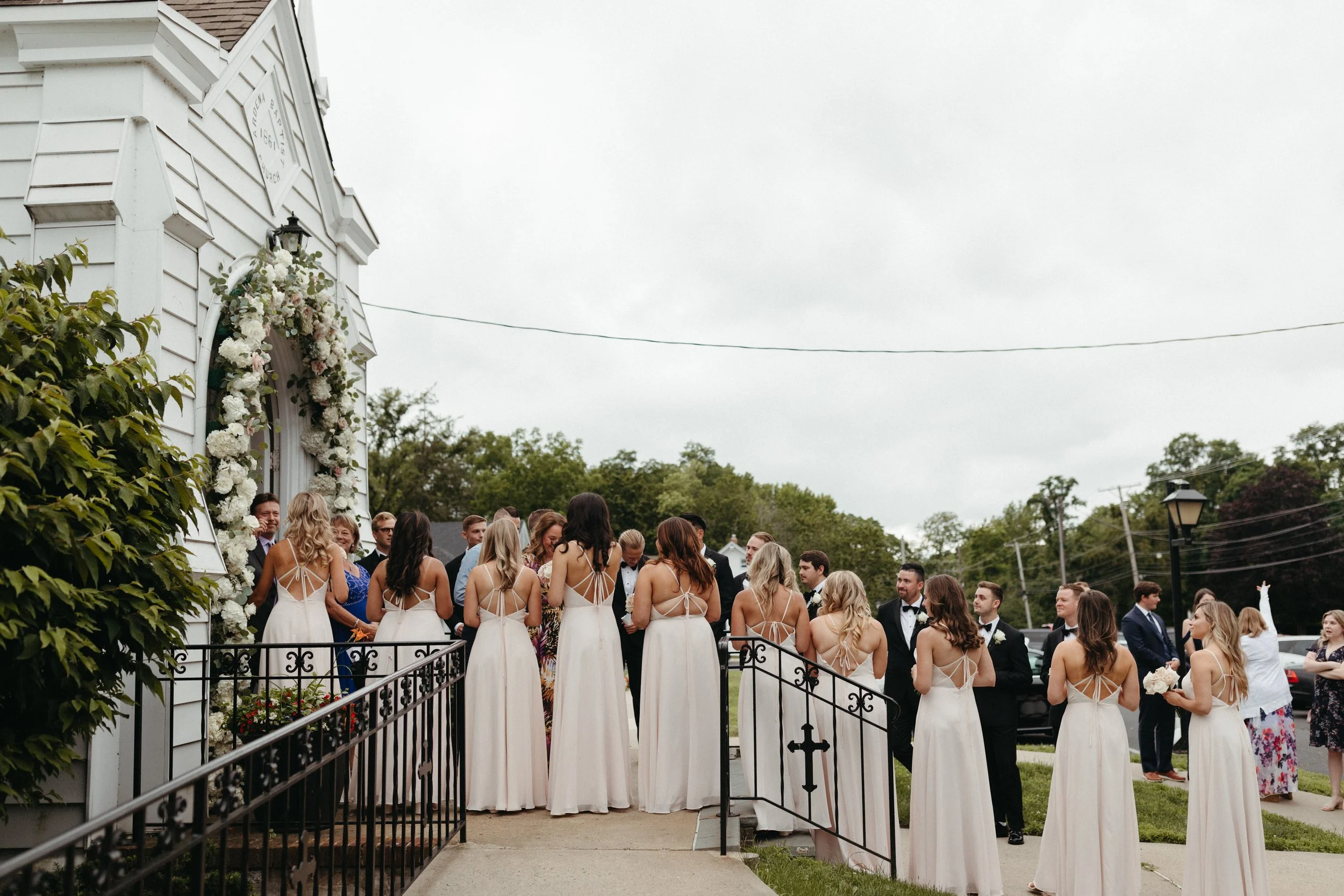 A wedding party gathered outside a white building with floral arrangements, with guests mingling and talking, under a cloudy sky.